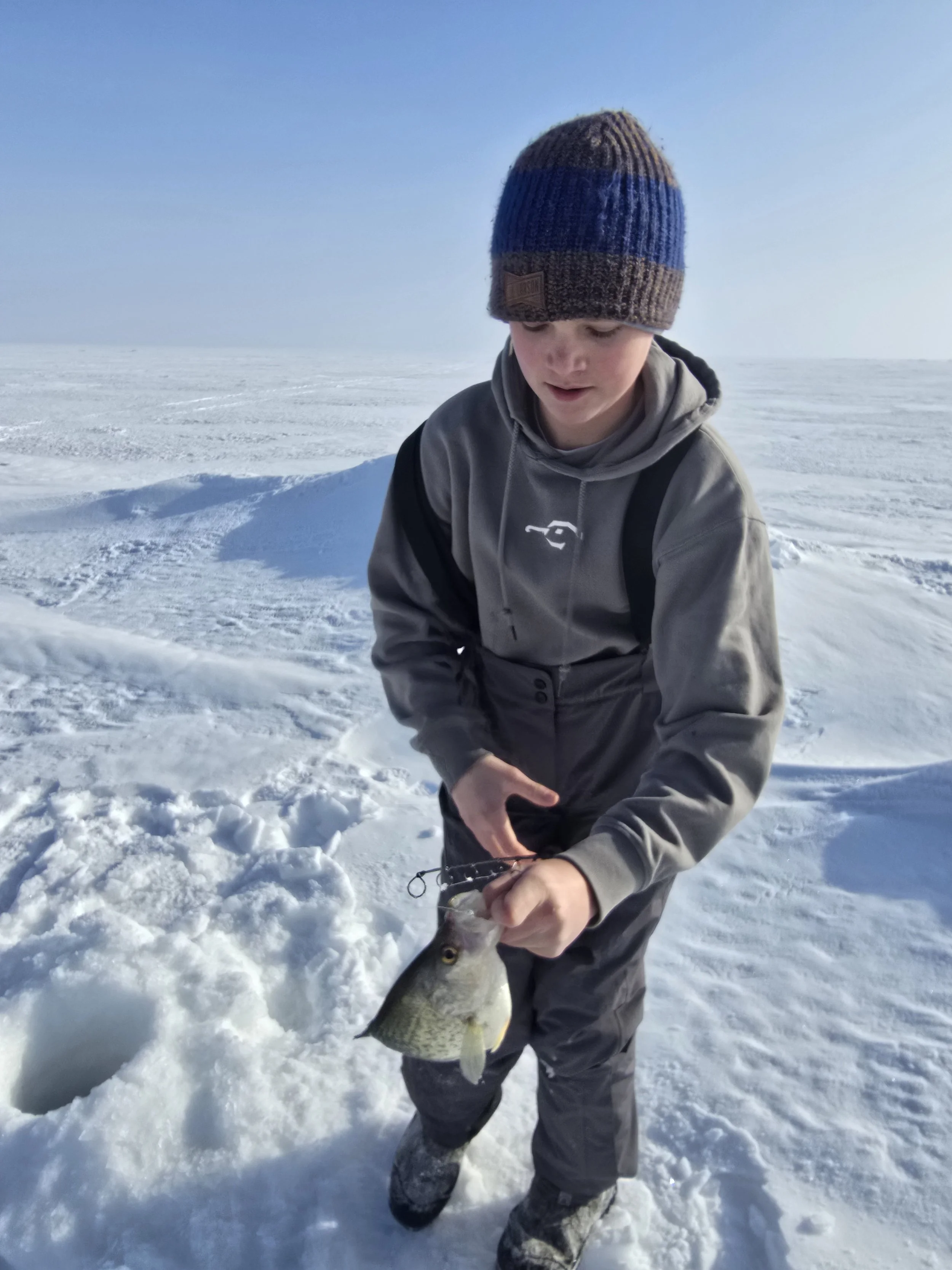 A young person wearing a blue and brown knit hat, gray jacket, and snow boots, standing on snow-covered ice in a cold, open landscape, holding a fish they caught.