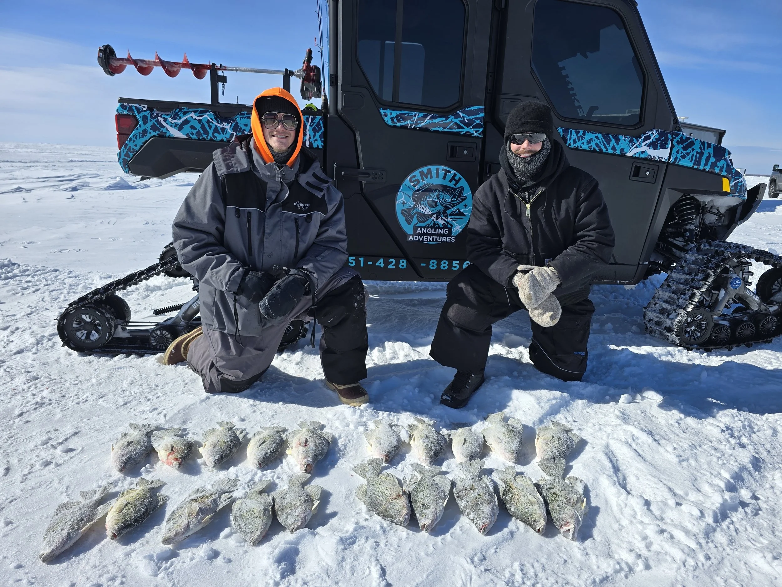 Two men kneeling in the snow with a collection of fish laid out in front of them and a snowmobile in the background. They are dressed warmly with sunglasses, winter jackets, and gloves, and appear to be on a frozen lake or snowy landscape.
