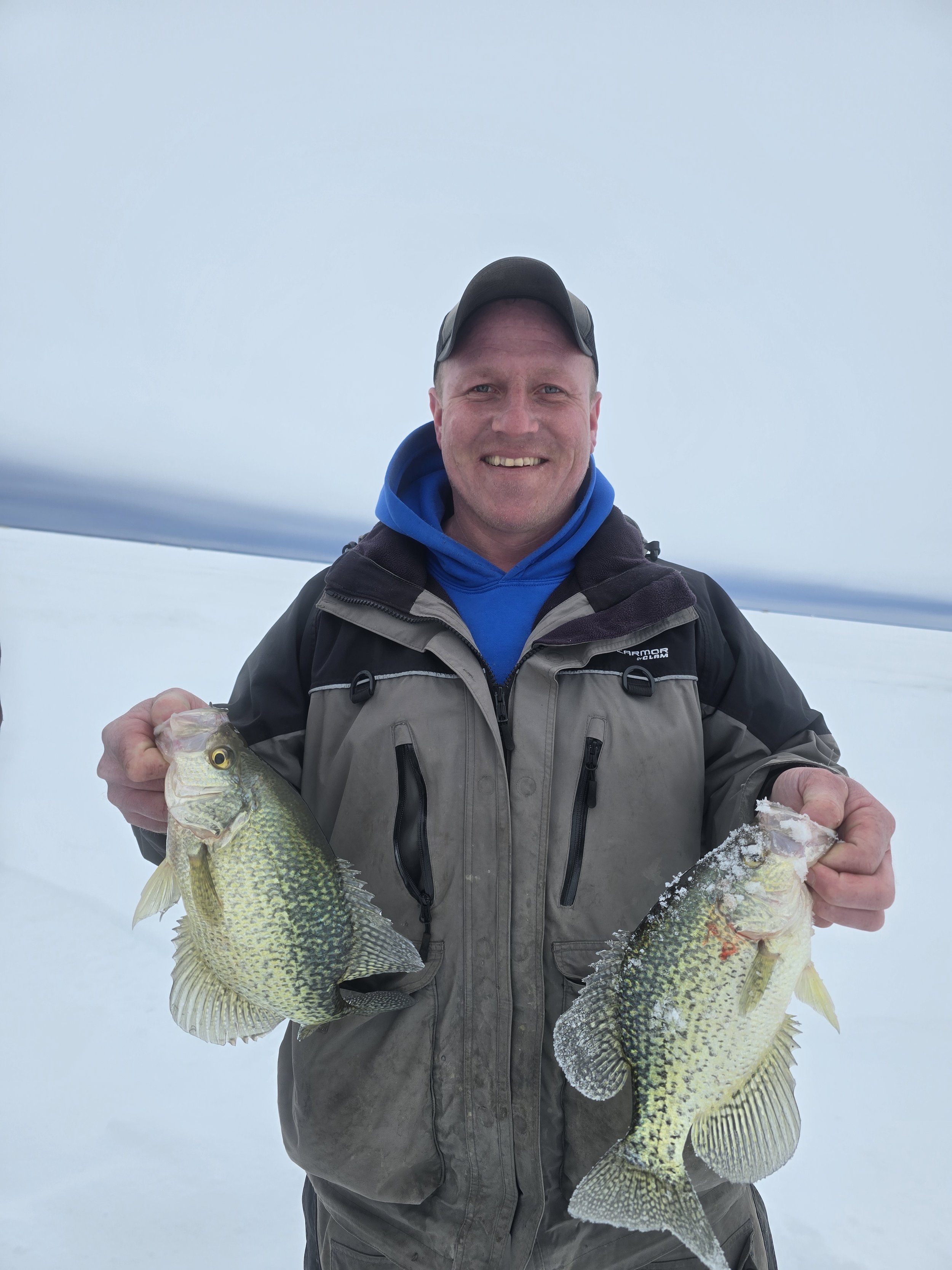 A man in outdoor winter gear holding two large fish, standing on a snowy landscape with an icy sky in the background.