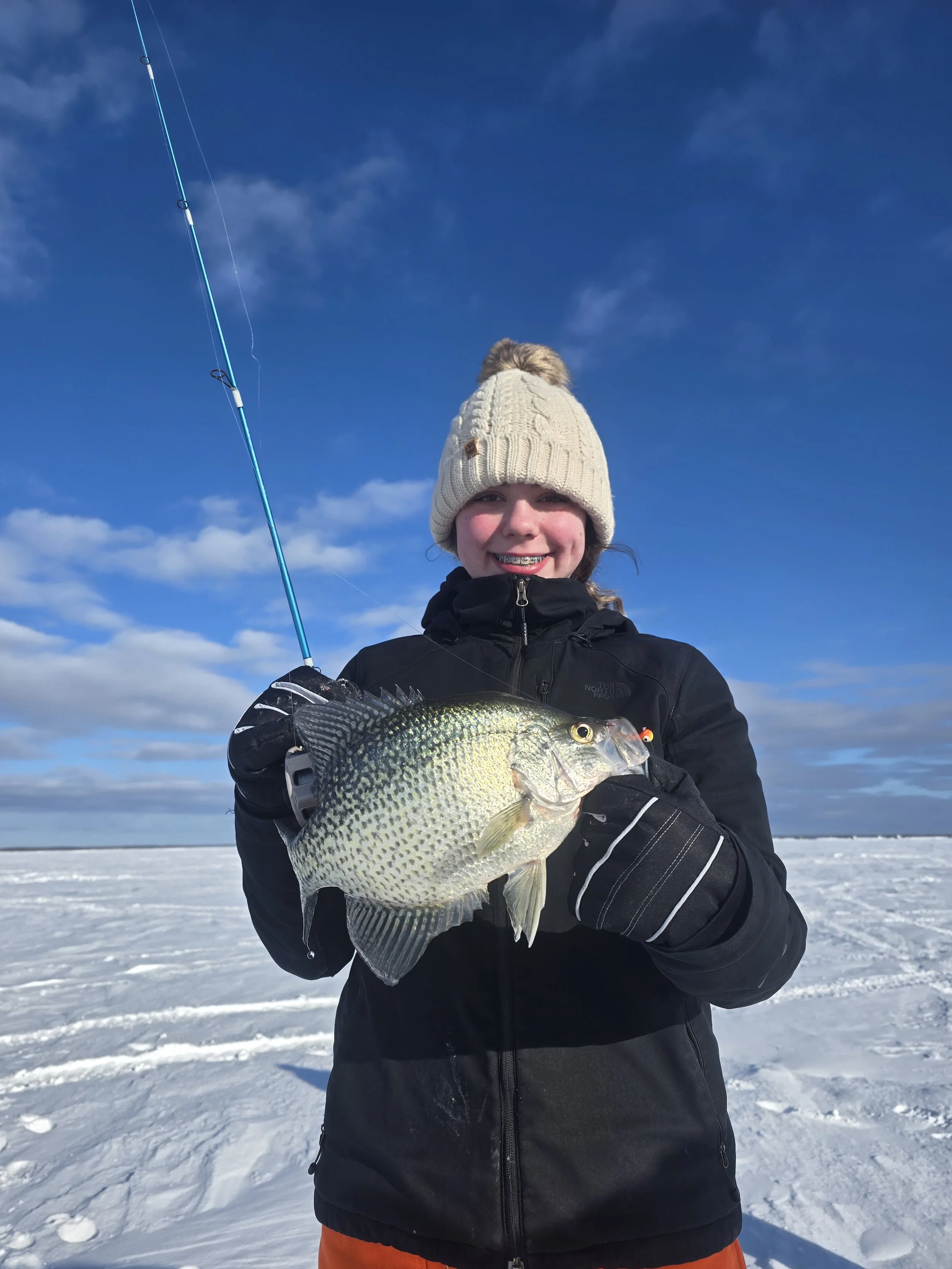 Young person outdoors in winter clothing holding a fish they caught, with a fishing rod in a snowy landscape under a partly cloudy sky.