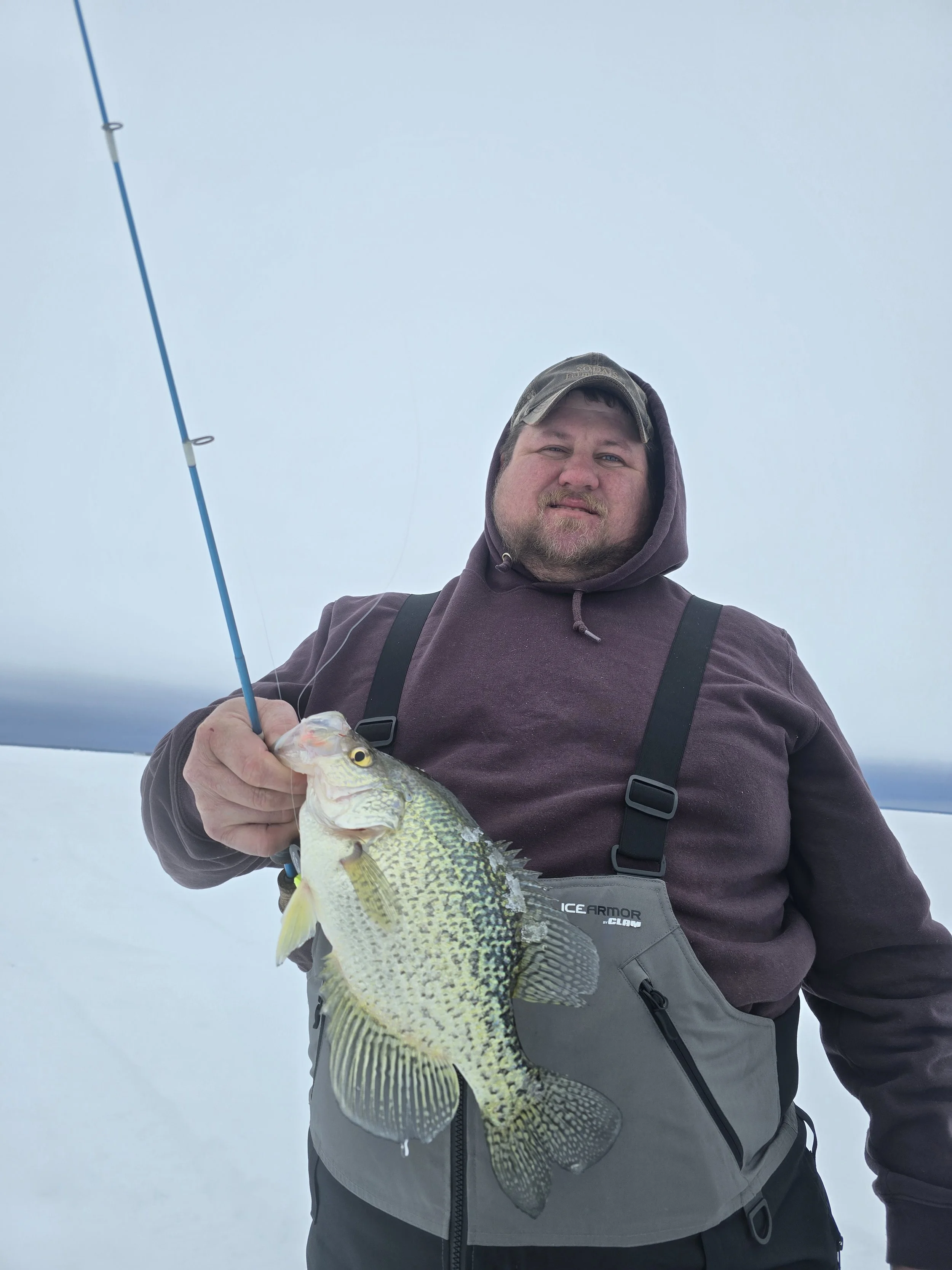 Man wearing a brown hoodie, hat, and overalls standing outdoors in snowy weather, holding a large fish with yellow and green markings.
