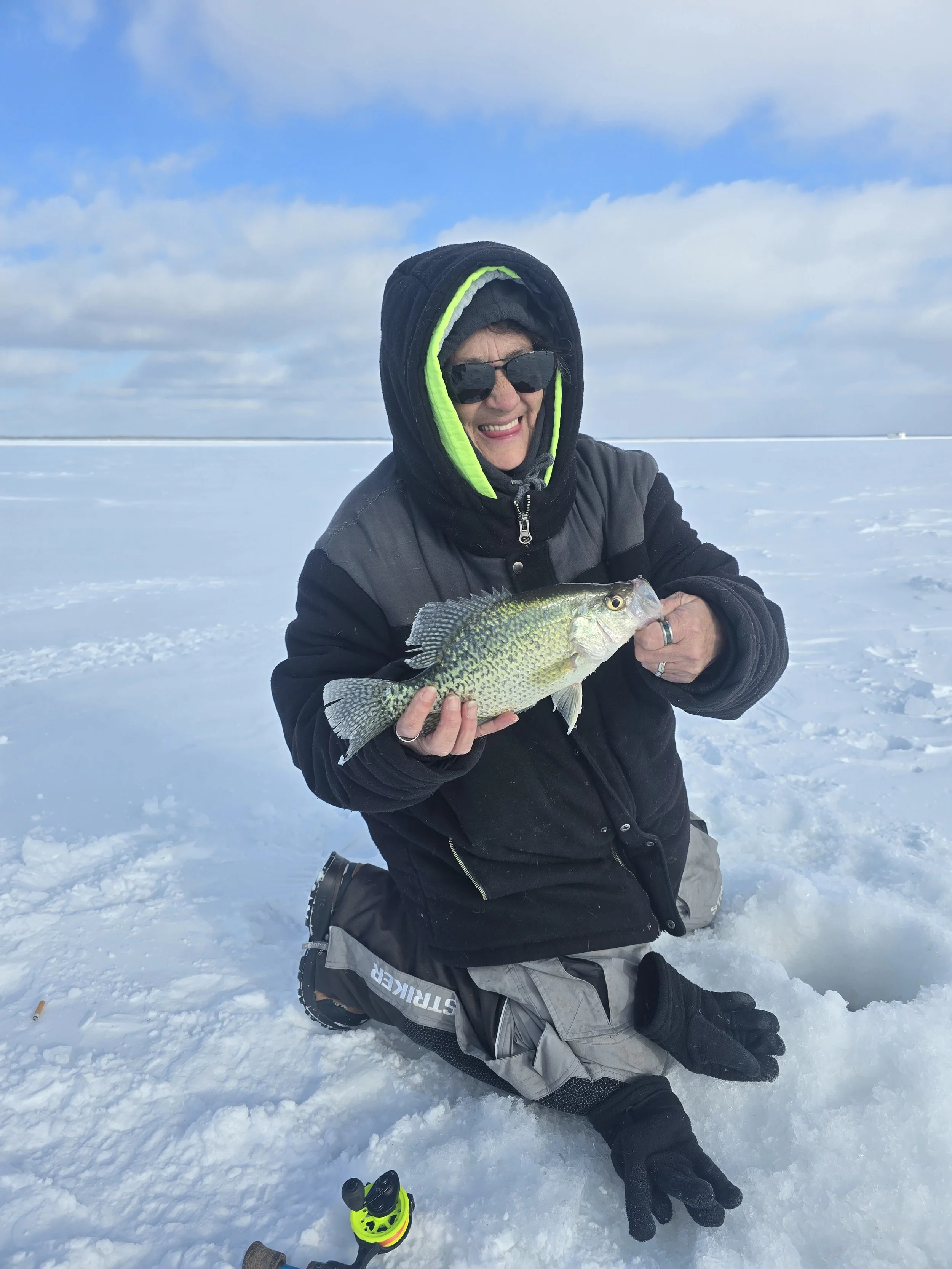 A person kneeling on snow holding a fish, dressed in winter clothing including a black hoodie with neon green lining, sunglasses, and a black jacket.