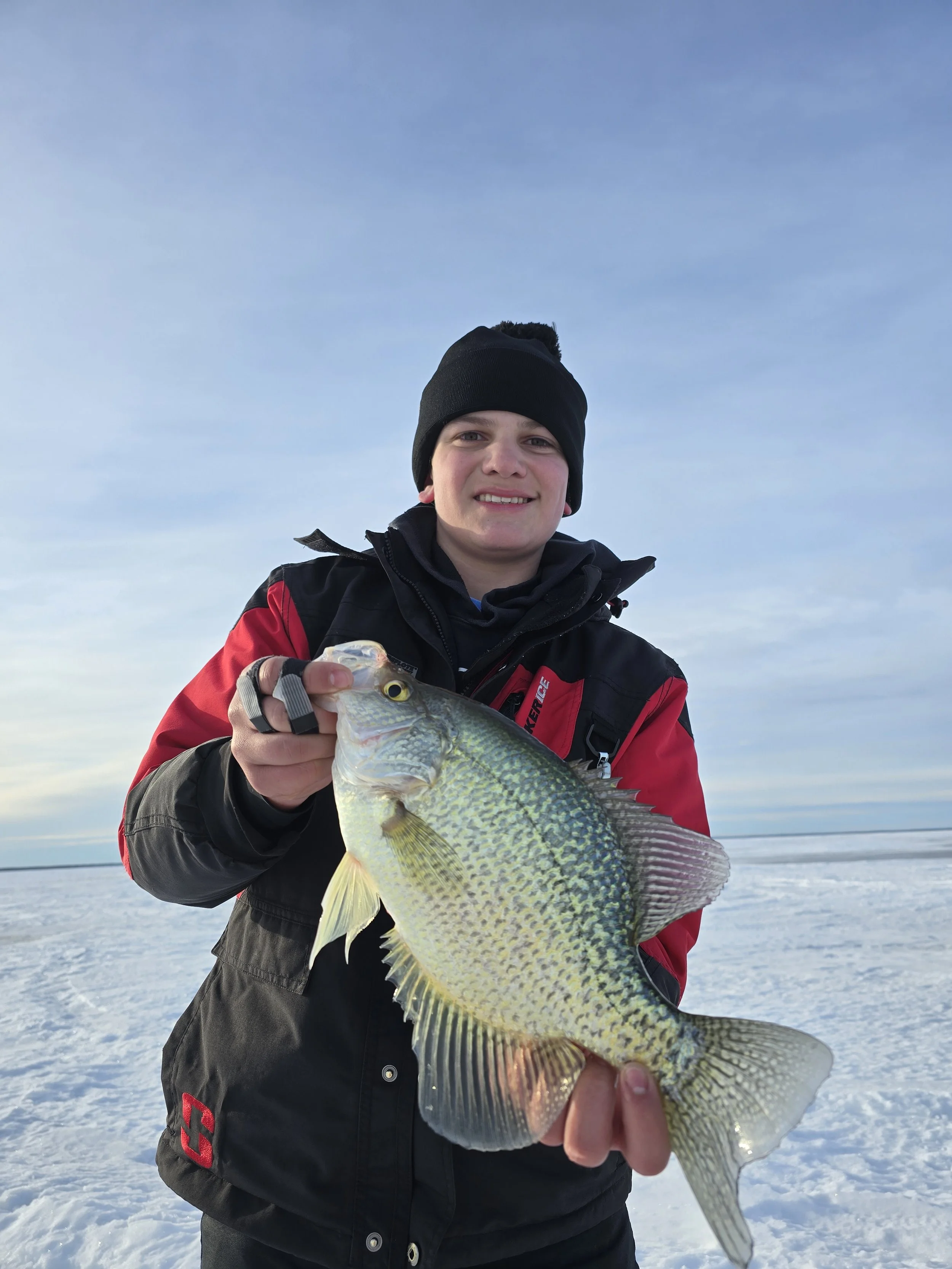 A young man outdoors on a snowy landscape, wearing a black beanie and red jacket, holding a large fish with yellow and green scales, smiling at the camera.