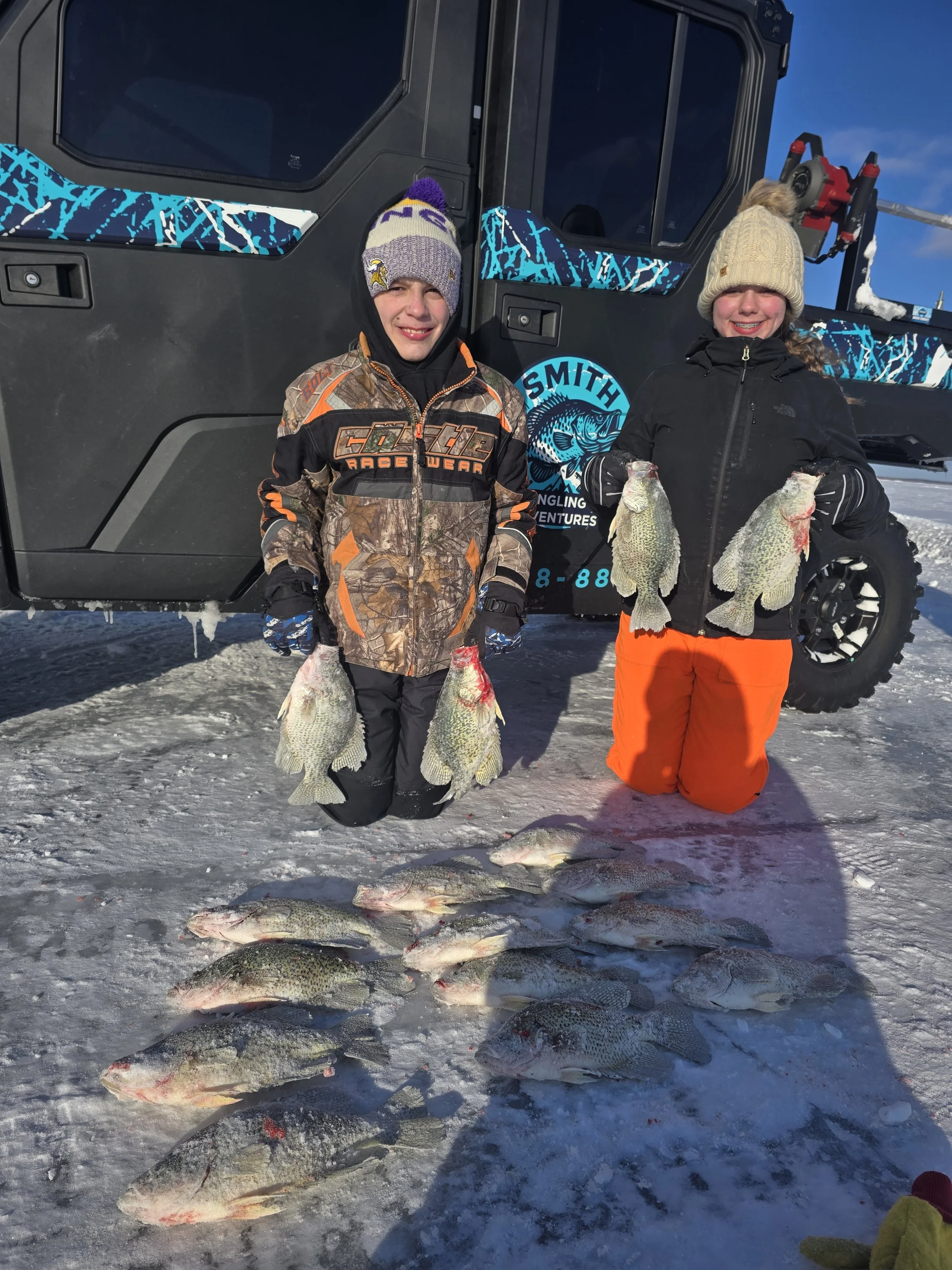 Two children in warm clothing stand on ice with fishing gear, holding fish they caught during a winter ice fishing adventure, with a snow-covered landscape and a vehicle in the background.