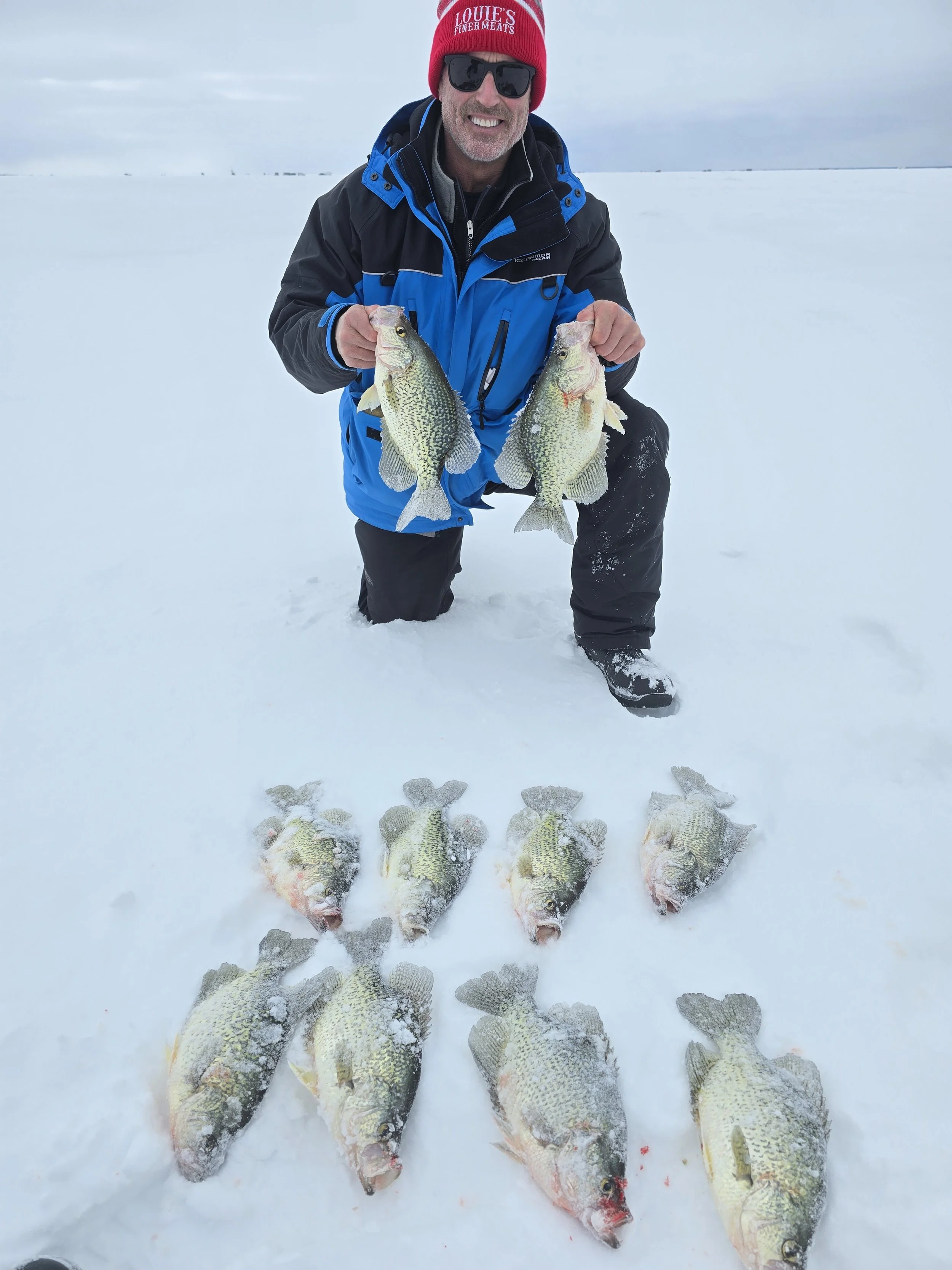 A man dressed in winter clothes, wearing sunglasses and a red hat that says "Louie's Finer Meats," is kneeling in the snow and holding two fish, with seven more fish laid out in front of him on the snow.
