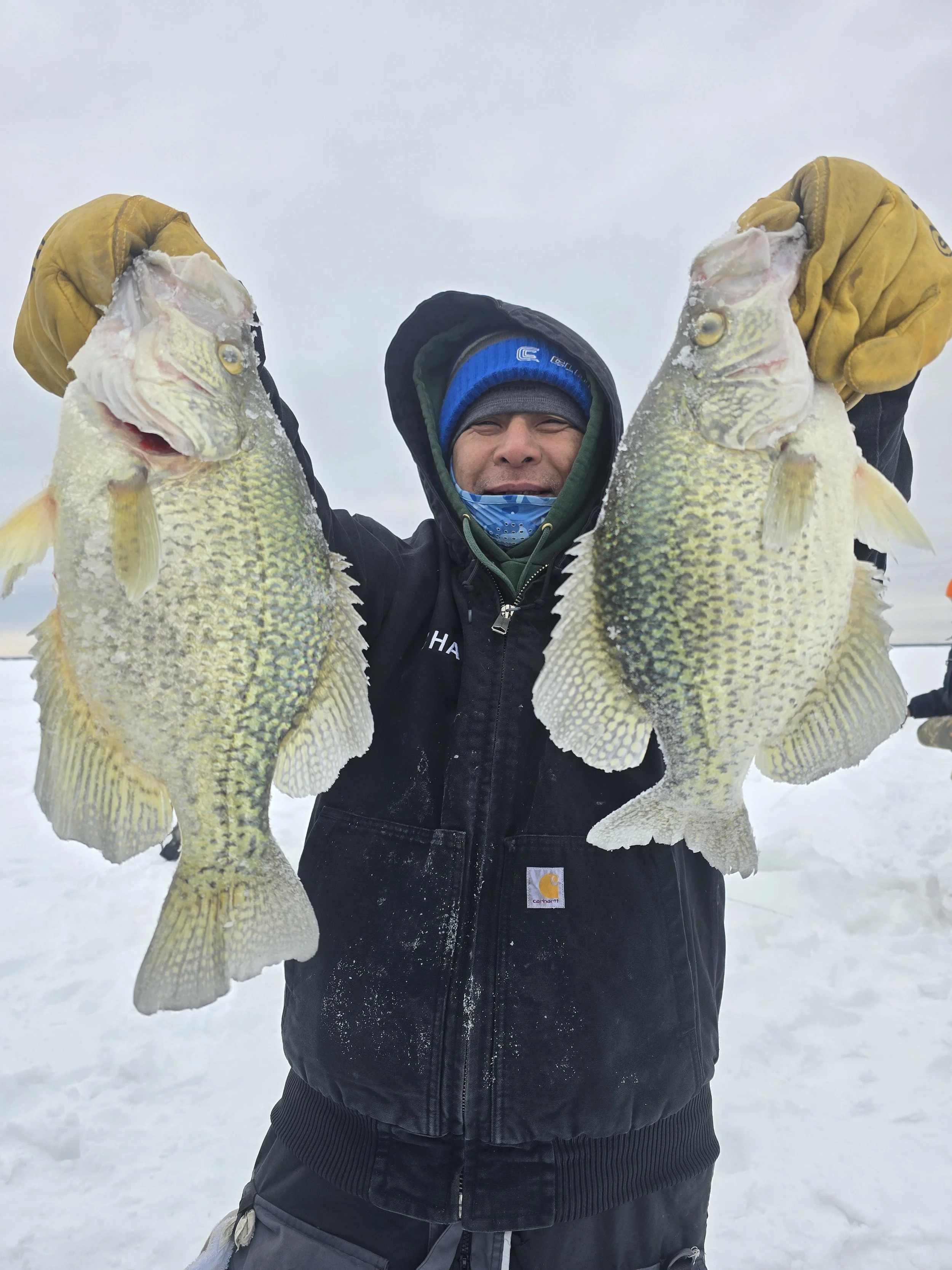 A man in winter clothing holding two large fish, standing on snow with an overcast sky.