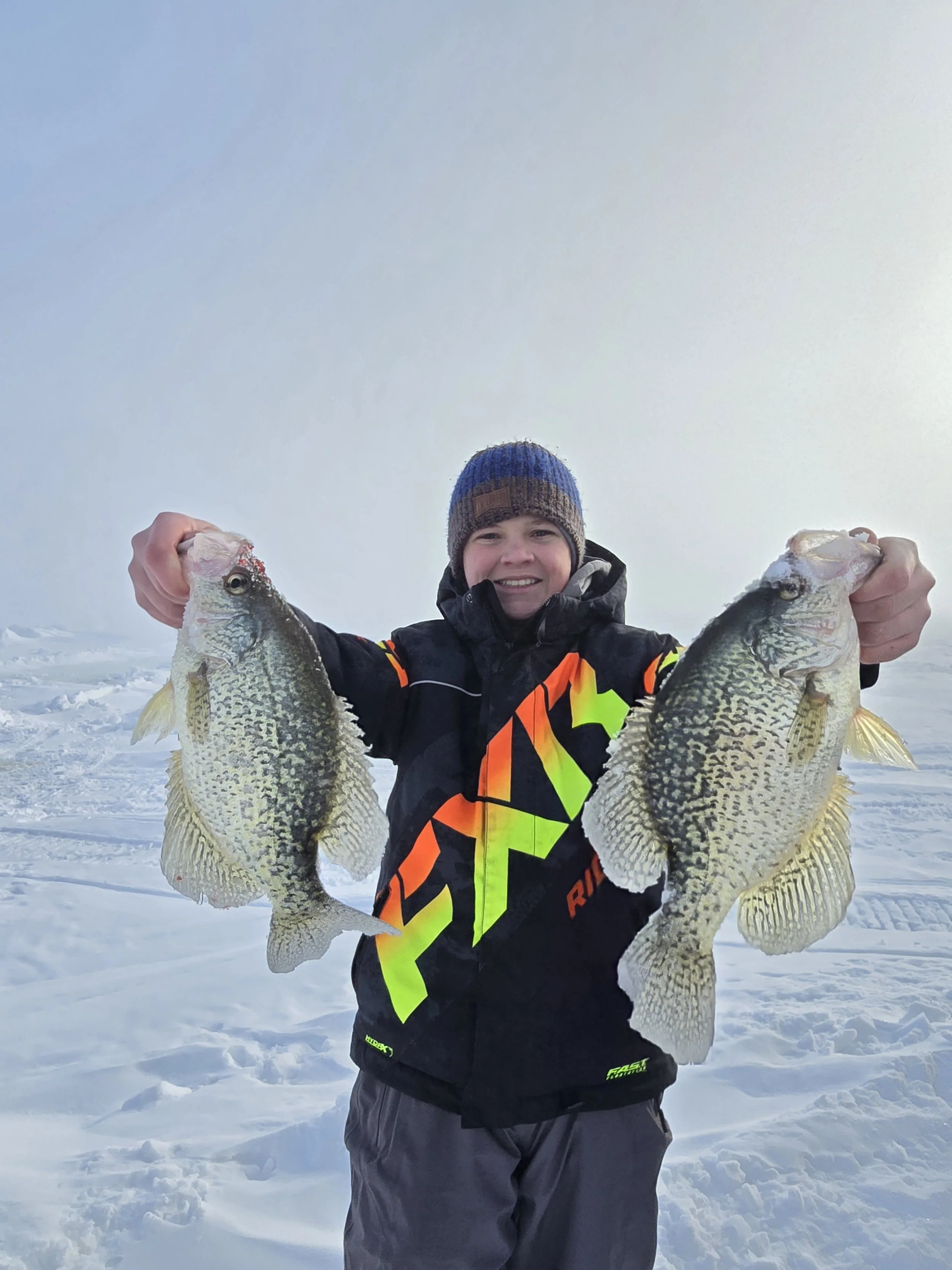 A smiling person dressed in winter clothing holding two large fish, standing on snow-covered ground, with a clear sky in the background.