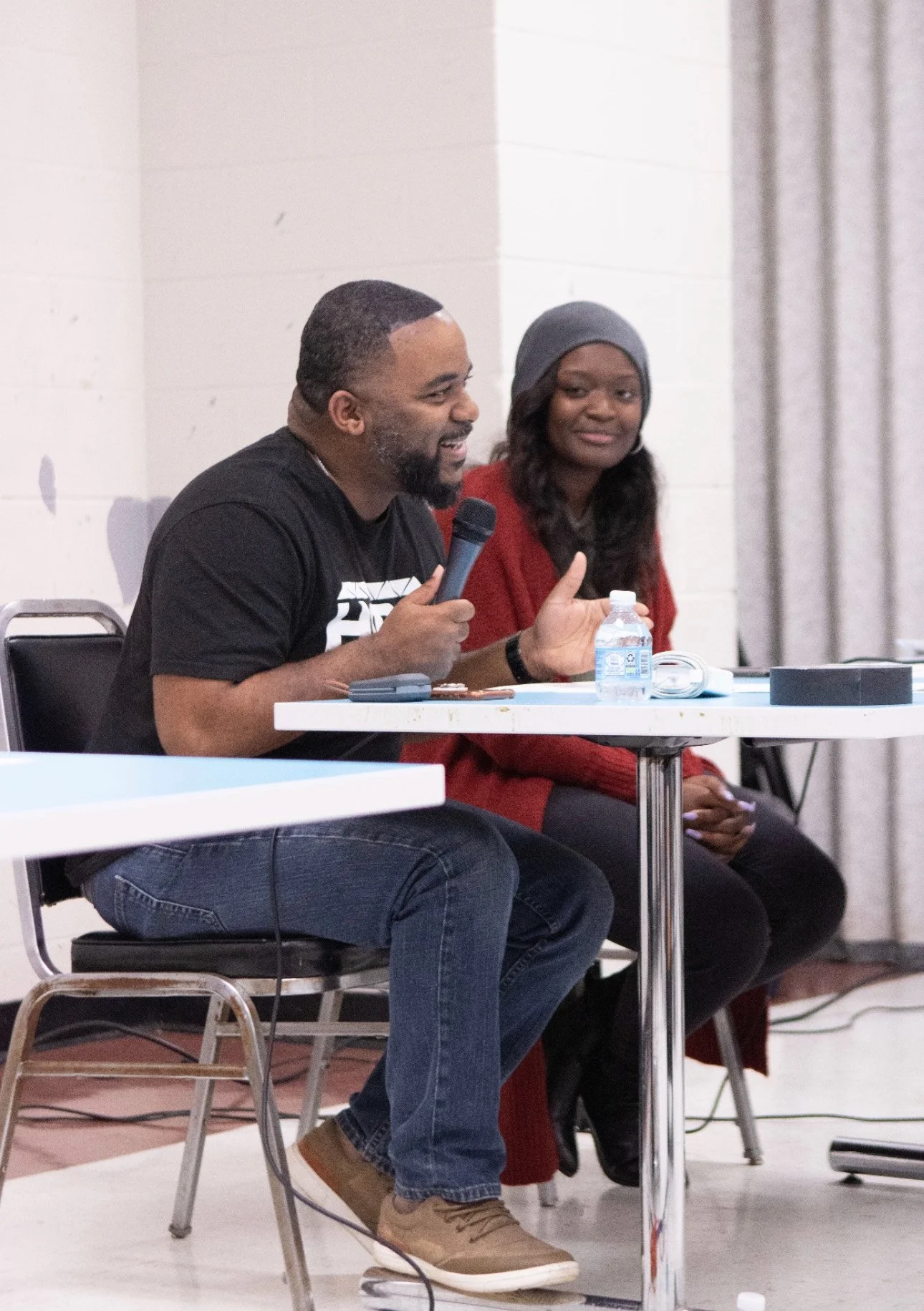 A man and a woman sitting at a table during a discussion or panel, with the man holding a microphone and smiling, and the woman looking on attentively.