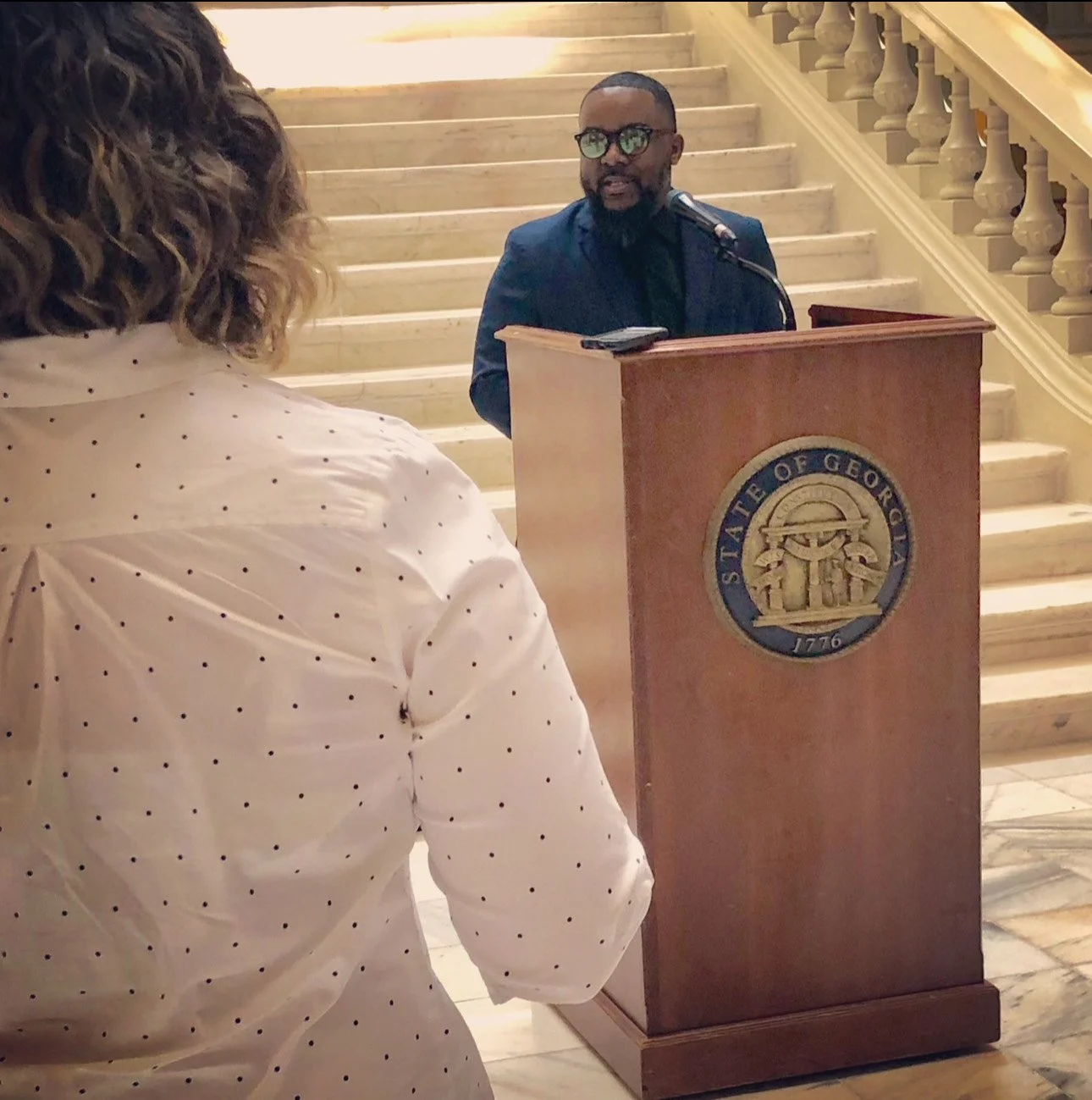 A man is speaking at a podium with the seal of the State of Georgia, in front of marble stairs in a formal setting, with a woman with curly hair and a white polka dot shirt in the foreground.