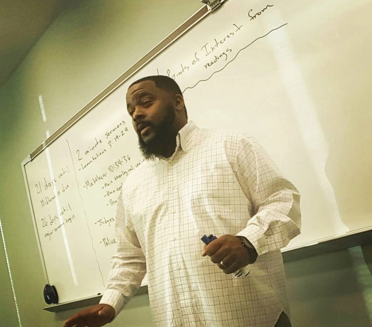 Man with dark skin, a beard, and short hair, standing in front of a whiteboard, holding a marker. The whiteboard has handwritten notes on it.