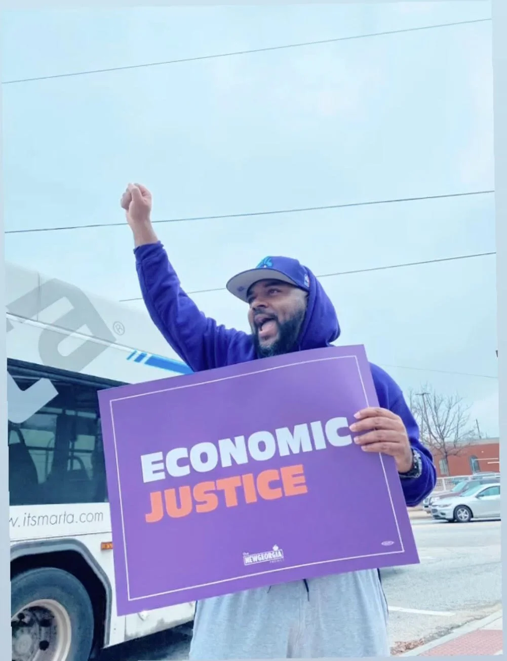 A man wearing a blue hoodie and cap holding a purple sign that says 'ECONOMIC JUSTICE,' raising his fist in the air during a protest or rally outside, with a bus and parked cars in the background.