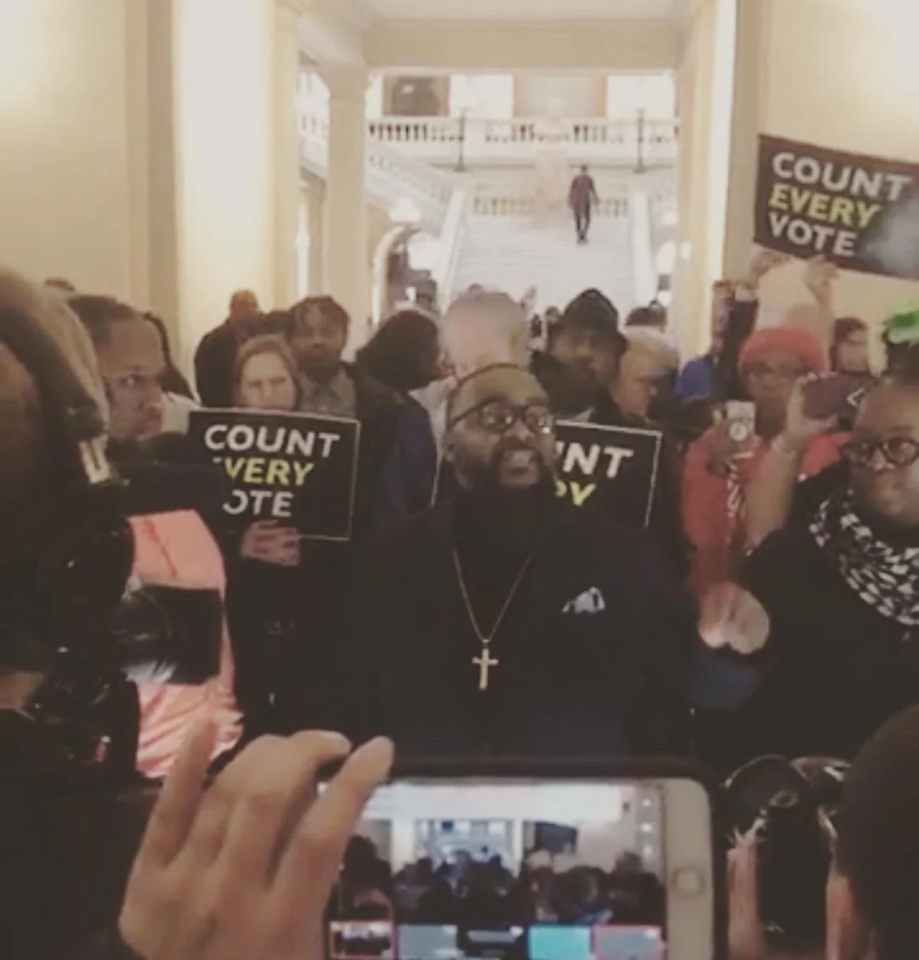 A group of people protesting and holding signs that say "Count Every Vote" inside a government building, with a staircase and a person walking down in the background.