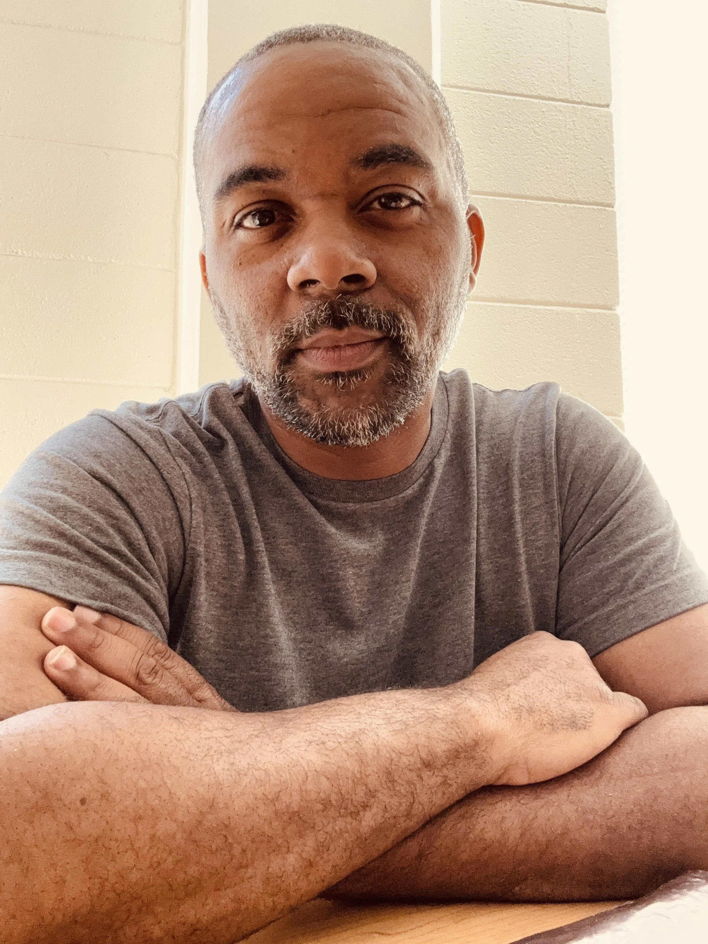 A man with a beard and mustache, wearing a gray T-shirt, sitting with arms crossed on a table against a light-colored wall.