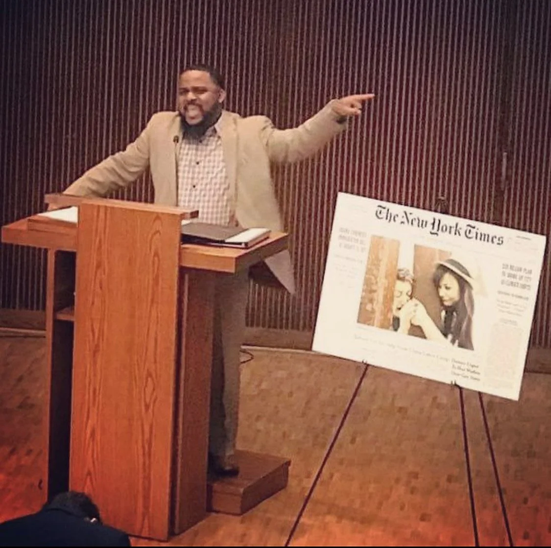 A man speaking at a wooden podium with follows news article featuring a photo of two women, one with curly hair and another with straight hair, on a stand beside him.