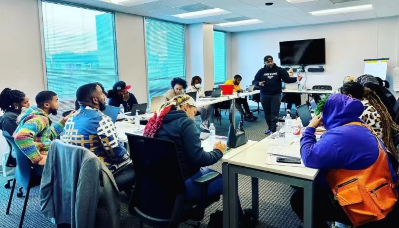 A diverse group of people attending a workshop or meeting in a conference room with large windows, seated around tables with laptops and notes, listening to an instructor standing near a TV screen.