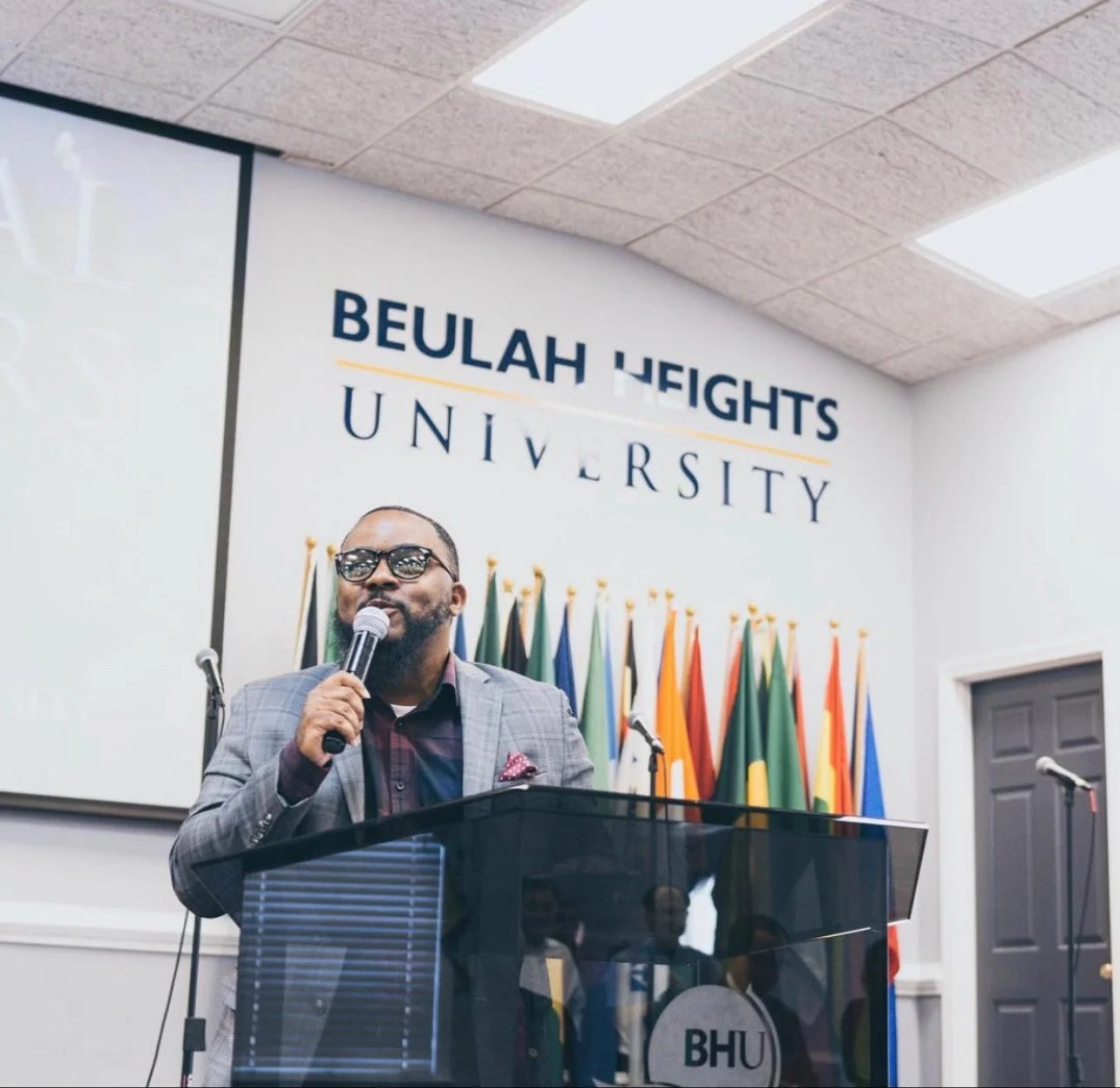 A man wearing glasses and a checkered blazer speaks into a microphone at a podium decorated with black glossy finish. Behind him, there are numerous international flags, and above, a sign reads 'BEULAH HEIGHTS UNIVERSITY' at Beulah Heights University
