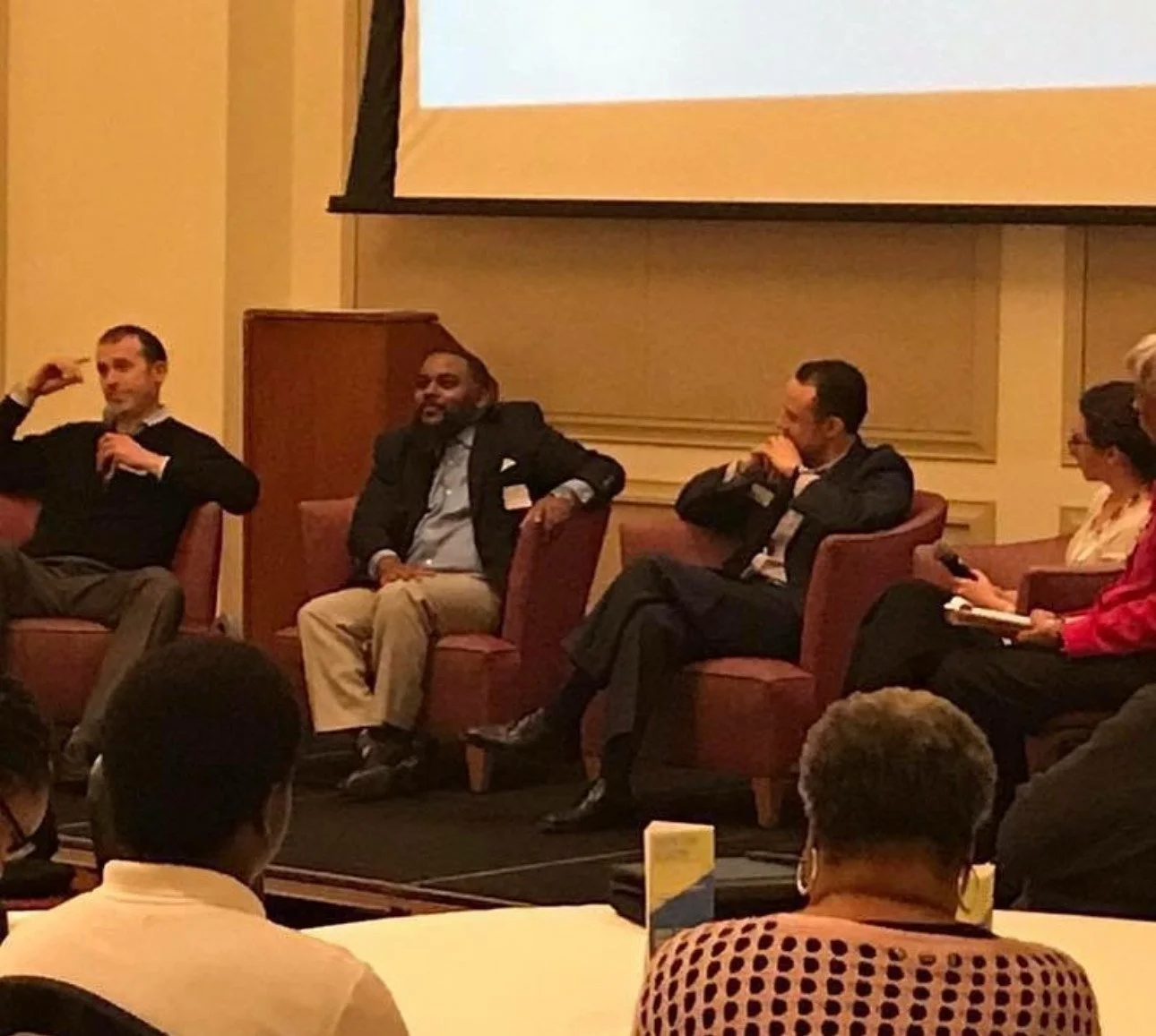 Panel discussion with five people seated on stage in a conference room. One person is speaking into a microphone, while others are listening or taking notes.