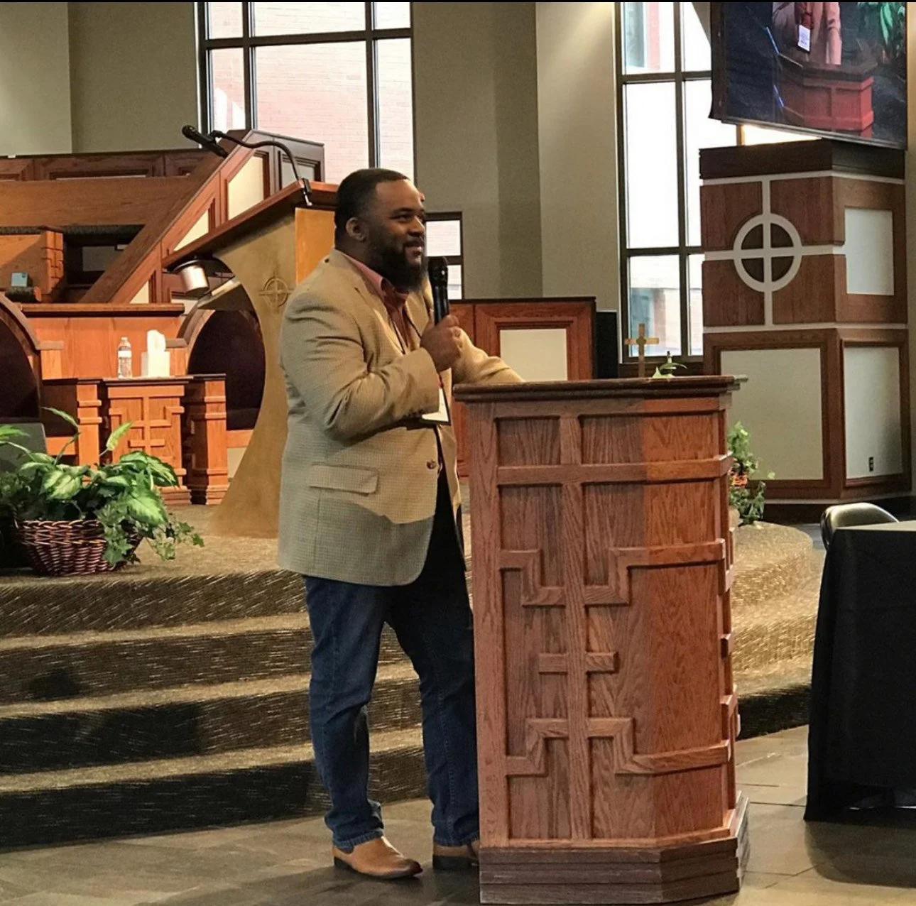 A man in a beige blazer and jeans stands at a wooden pulpit with a cross carved into the front, speaking into a microphone inside a church or chapel. Behind him are large windows, a pulpit, and a projection screen.