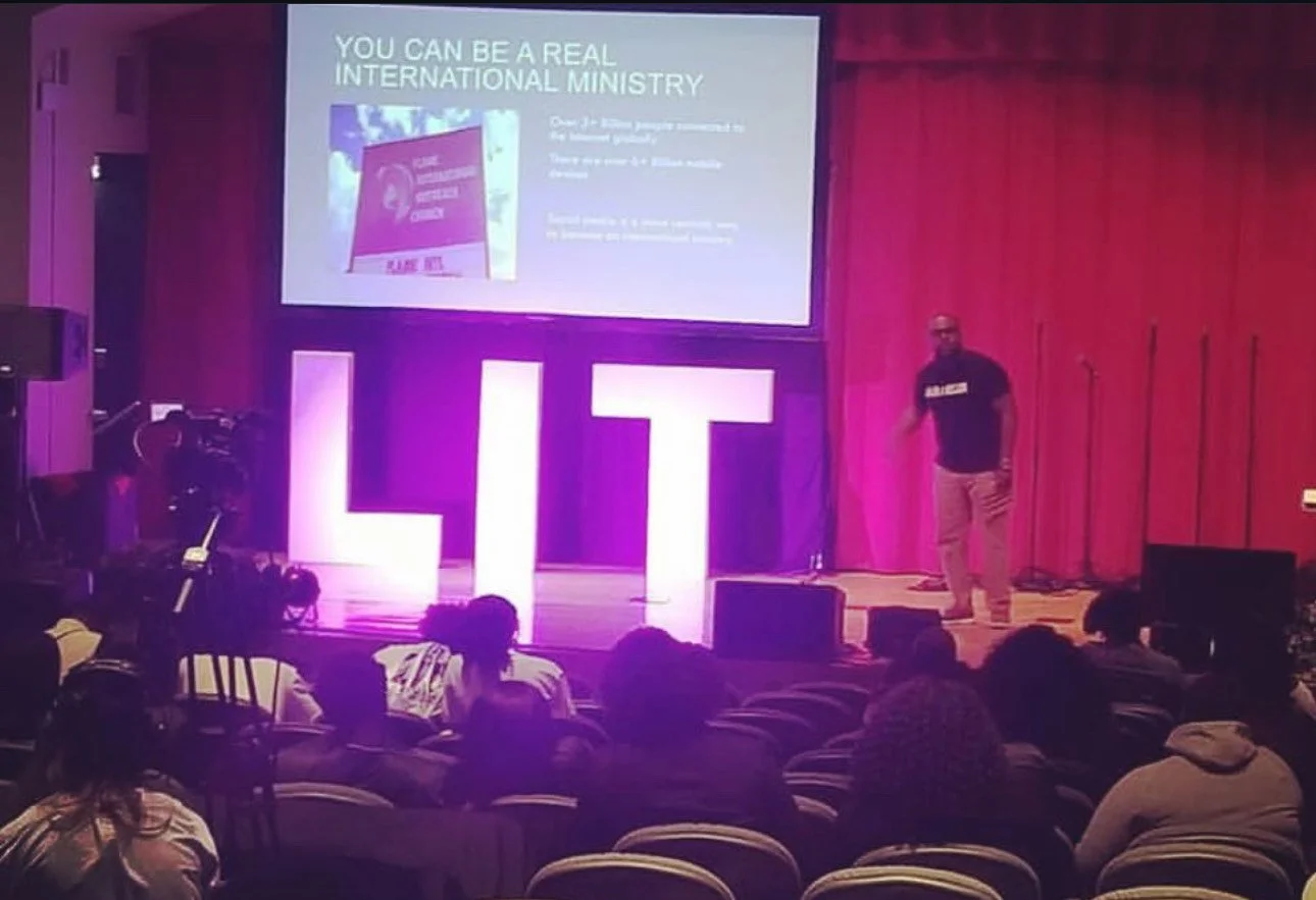 A man standing on a stage with large illuminated letters spelling 'LIT' in front of him, giving a presentation about how to be a real international ministry. The audience sits in chairs facing the stage, and there is a large screen displaying a slide