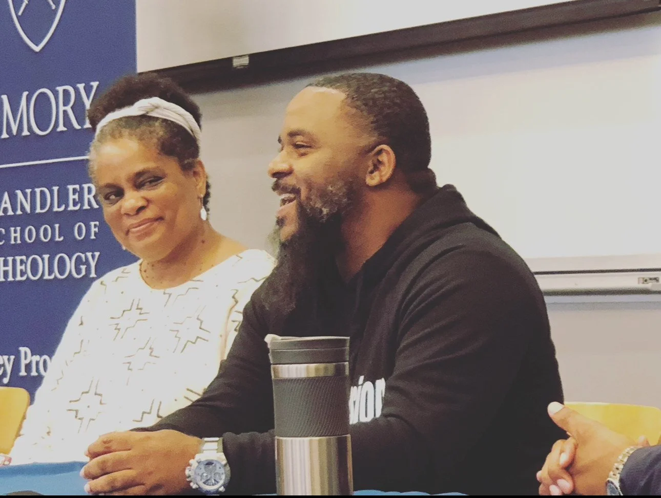 Two people speaking at a panel discussion at a university event, with a woman wearing a white dress and a man wearing a black hoodie, seated in front of a blue banner with the university's name.