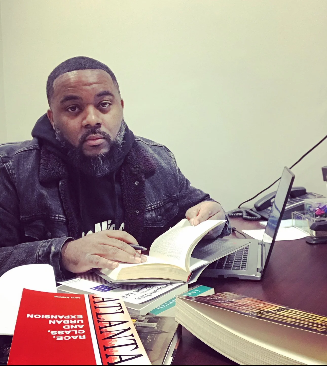 A man sitting at a desk with an open book, a laptop, and several other books. He has a dark hoodie and a black jacket, with a beard and short hair, looking at the camera.