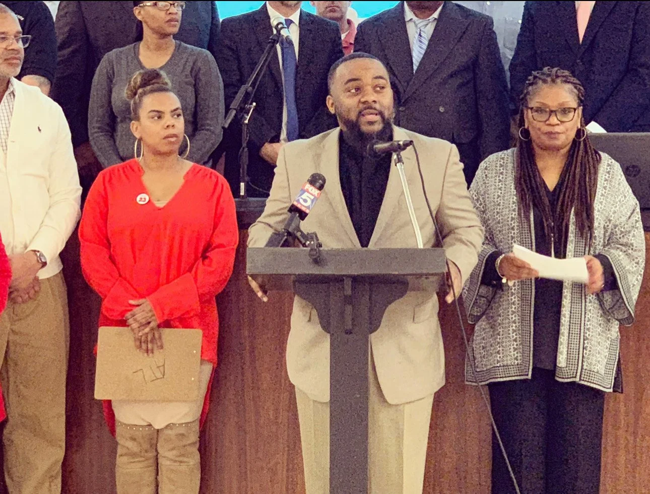 A man in a light gray suit speaking at a podium during a press conference, with a microphone, flanked by two women, one in a red outfit holding a folder, and the other in glasses holding papers, surrounded by a group of people in formal attire.