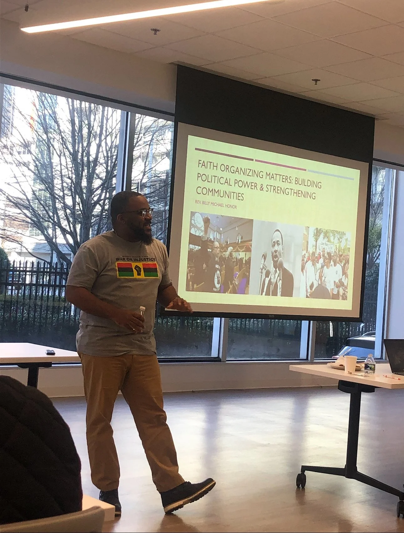 A man giving a presentation on faith organizing and community building, standing beside a large screen with a slide titled 'Faith Organizing Matters: Building Political Power & Strengthening Communities' in a room with large windows and trees outside