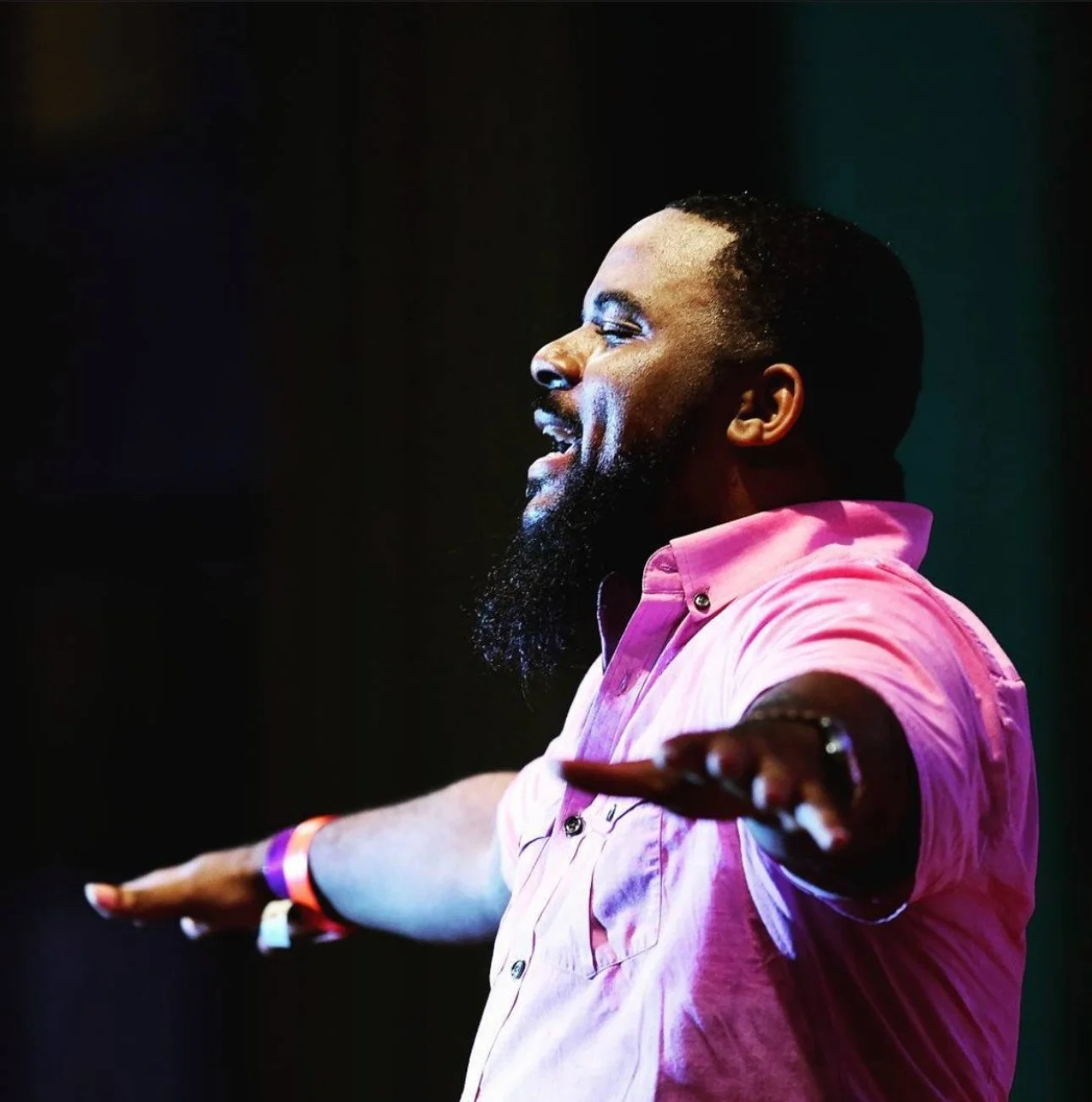 A man with a beard and short hair wearing a pink shirt, standing in profile with arms outstretched, smiling, against a dark background.