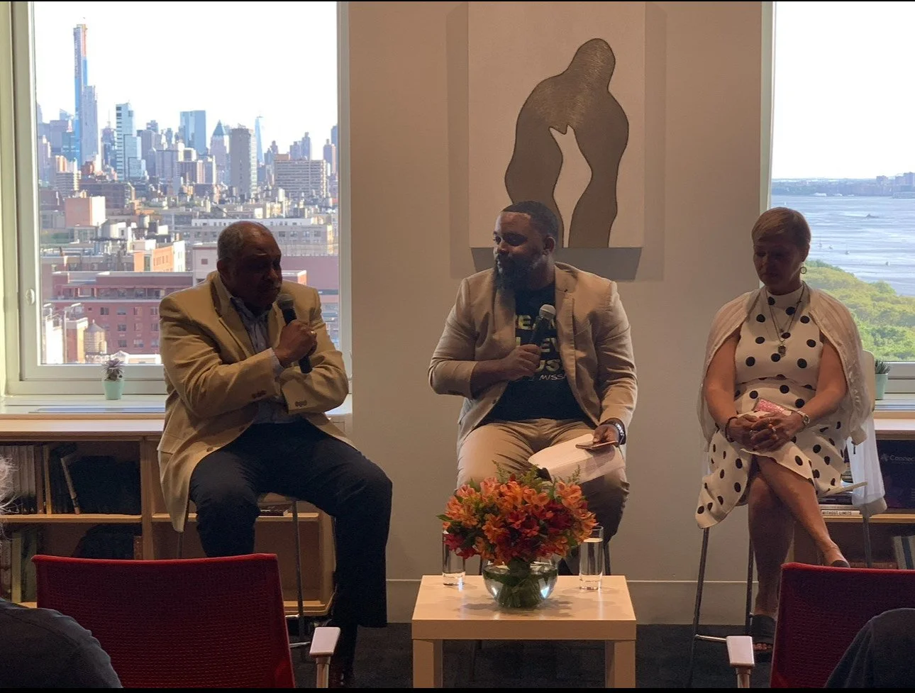 Three people sitting in a panel discussion, each holding a microphone, with city skyline and water view through windows behind them, and a painting of two embracing figures on the wall.