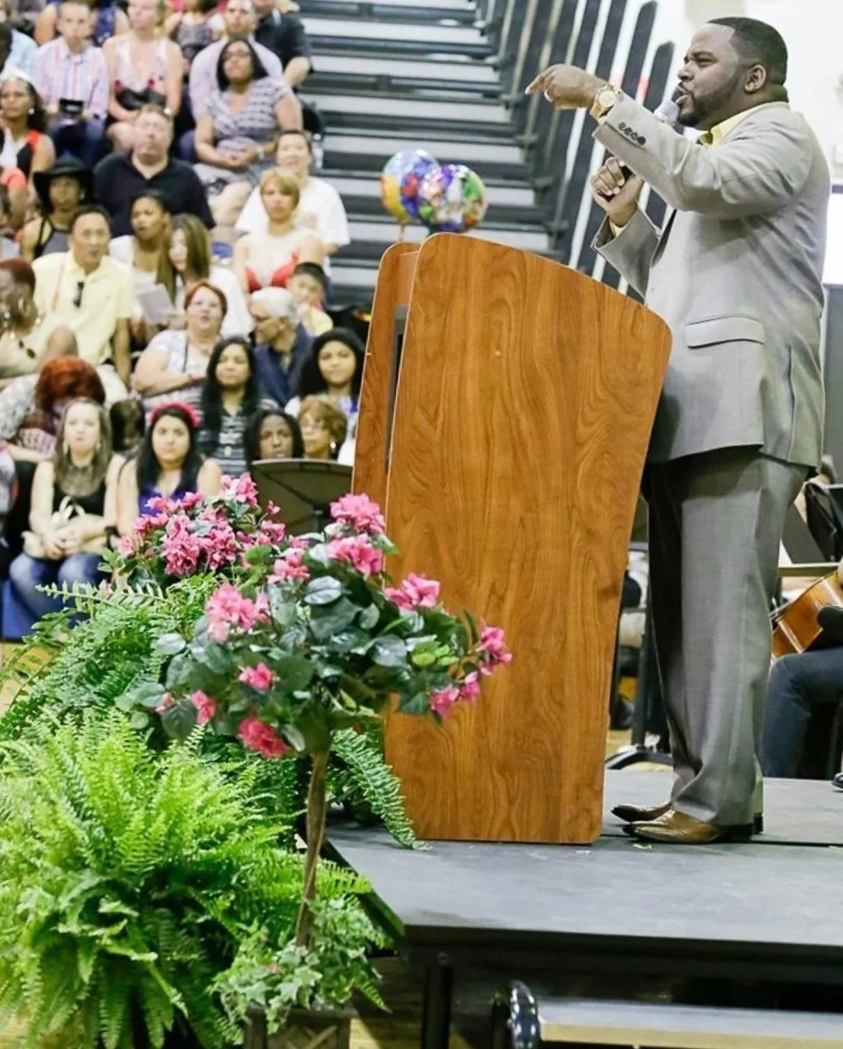 A man in a gray suit speaking at a podium decorated with pink flowers, addressing a diverse audience seated in an auditorium.