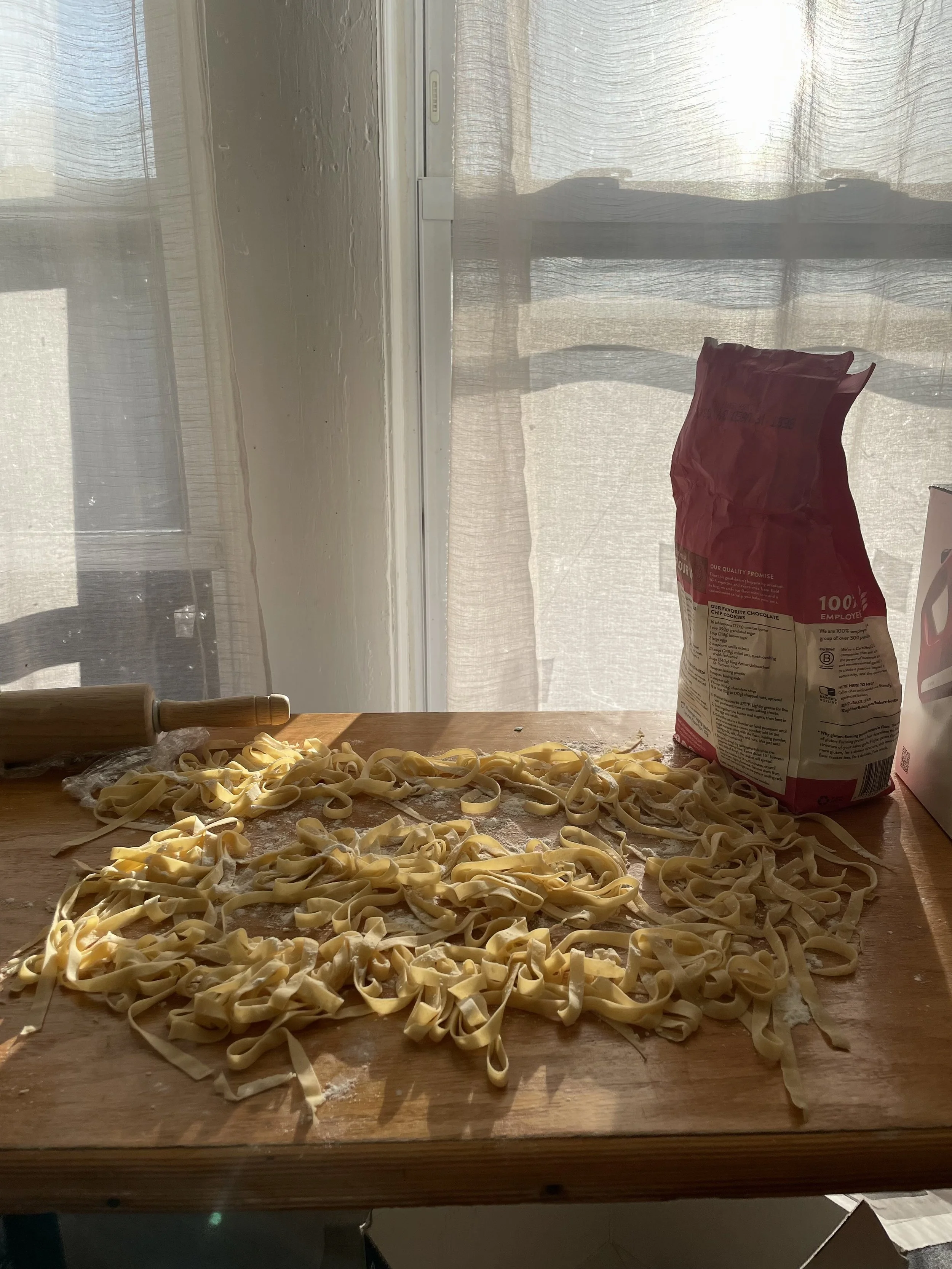 Fresh homemade pasta noodles on a floured wooden countertop with a rolling pin and a bag of flour nearby, in front of a window with sheer curtains.