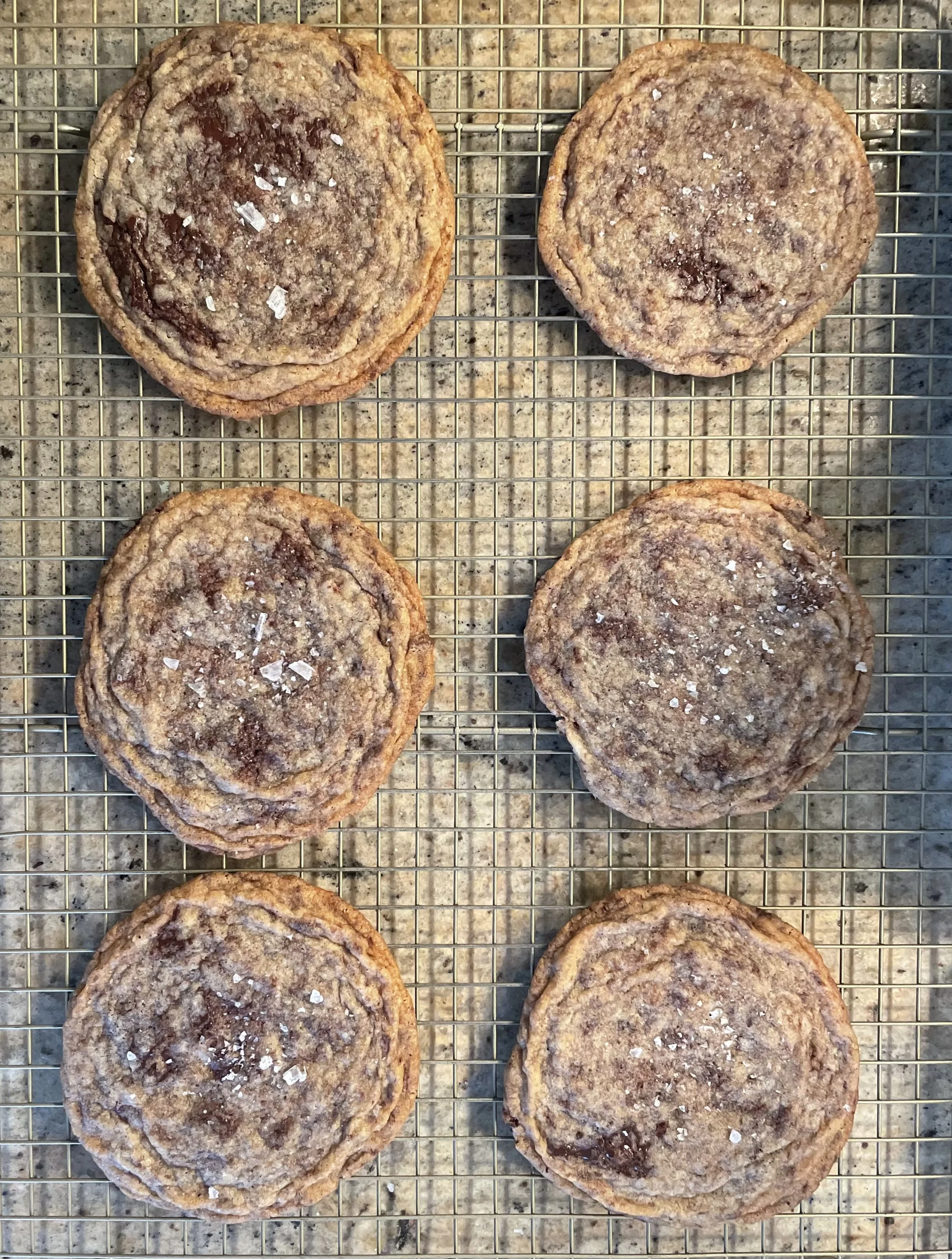 Six chocolate chip cookies with salt crystals on top, arranged in two rows on a wire cooling rack.