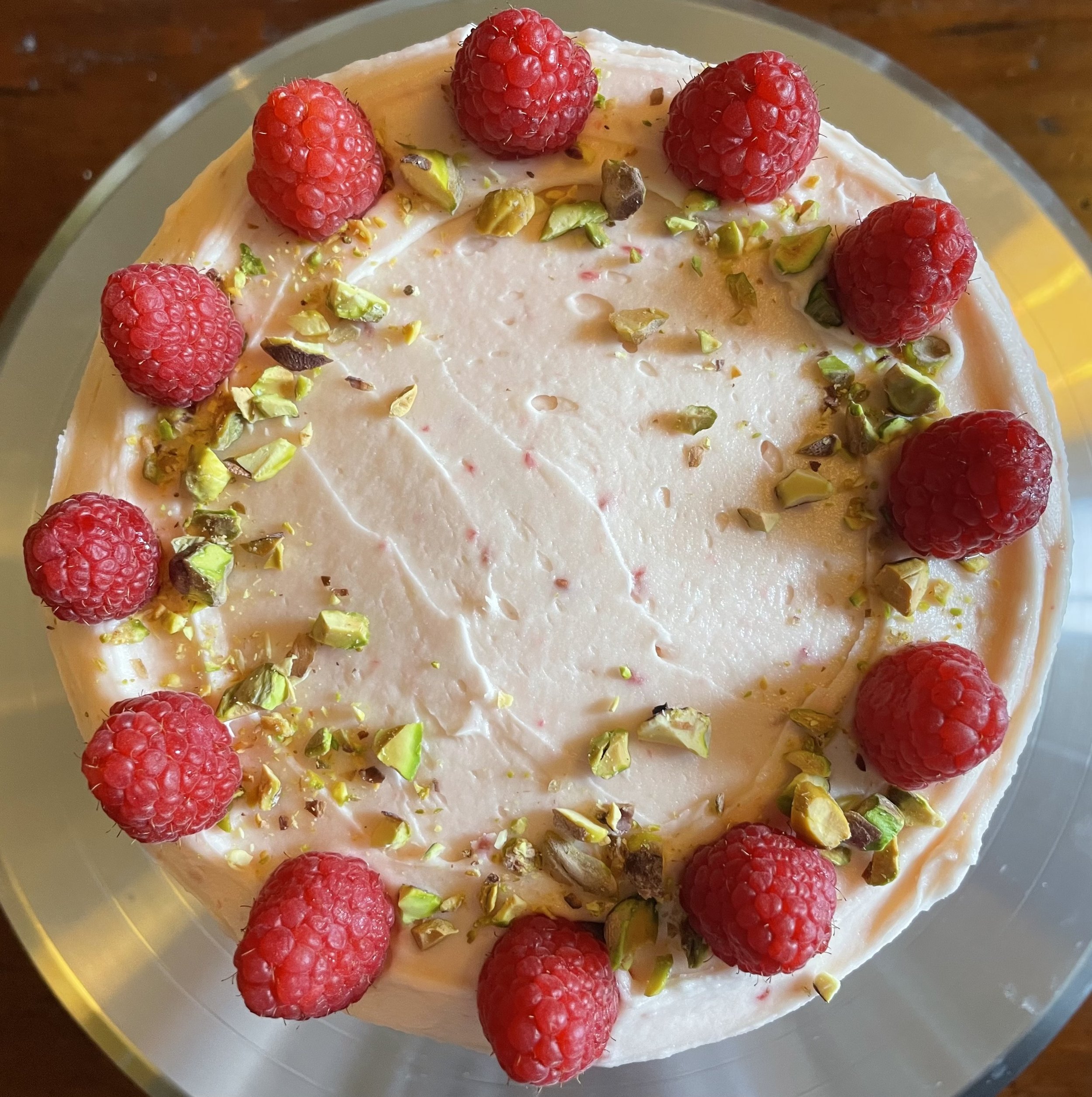 A round cake decorated with fresh raspberries and chopped pistachios on top.