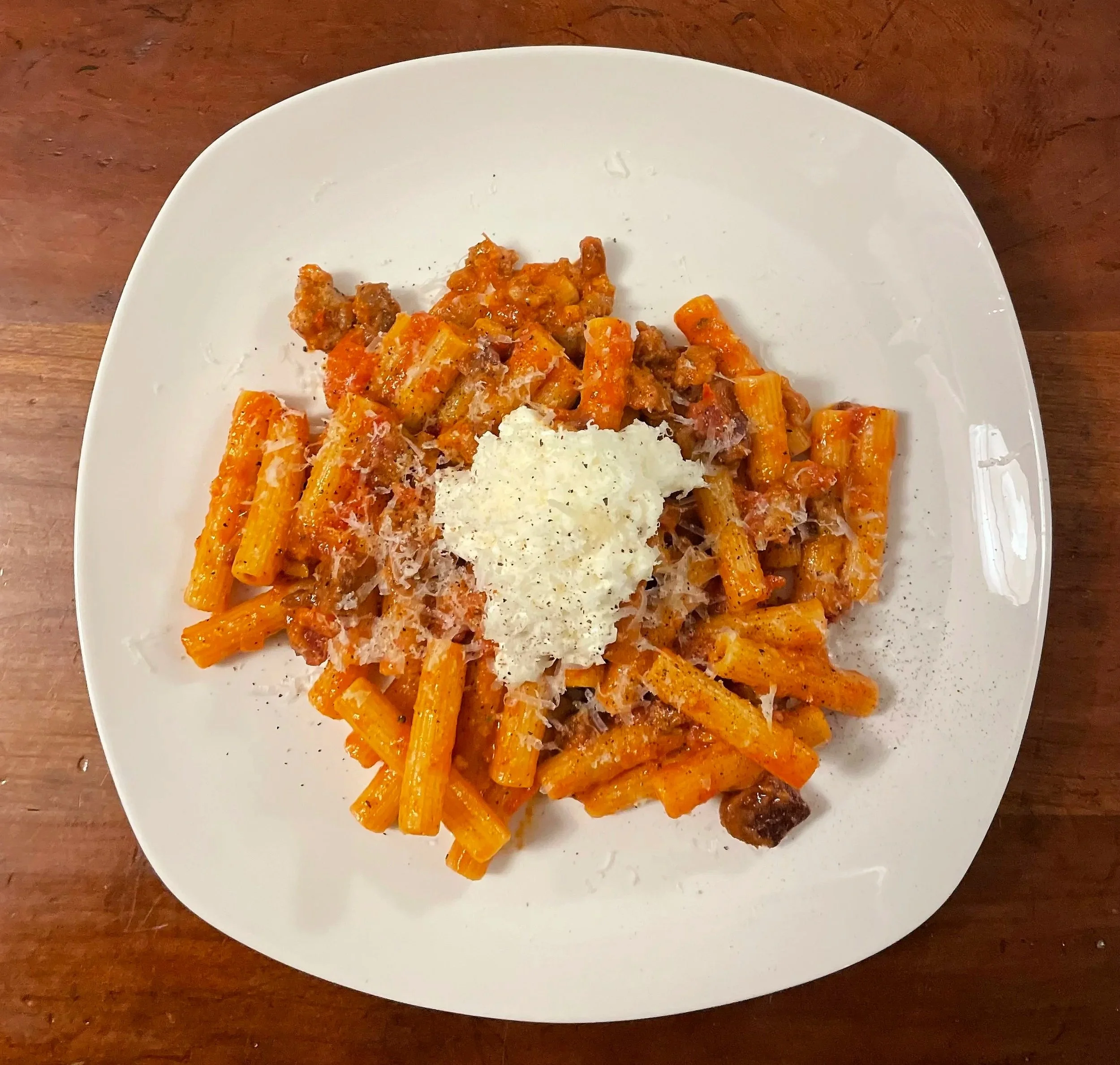 A white plate with cooked rigatoni pasta topped with tomato sauce, grated cheese, and crumbled sausage, on a wooden table.