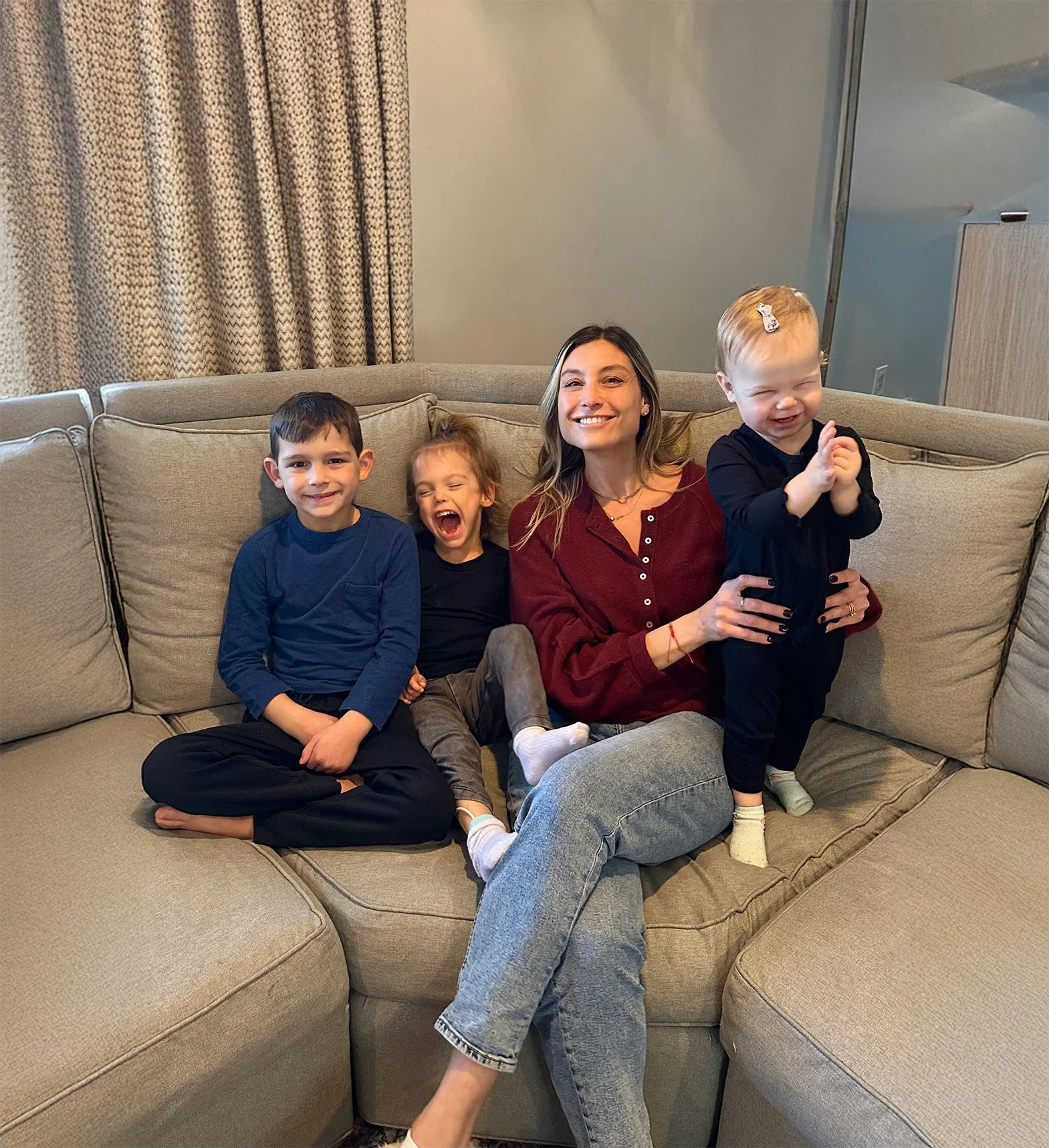 A woman and three young children sitting on a beige sectional sofa, smiling and laughing in a living room with gray walls and curtains.