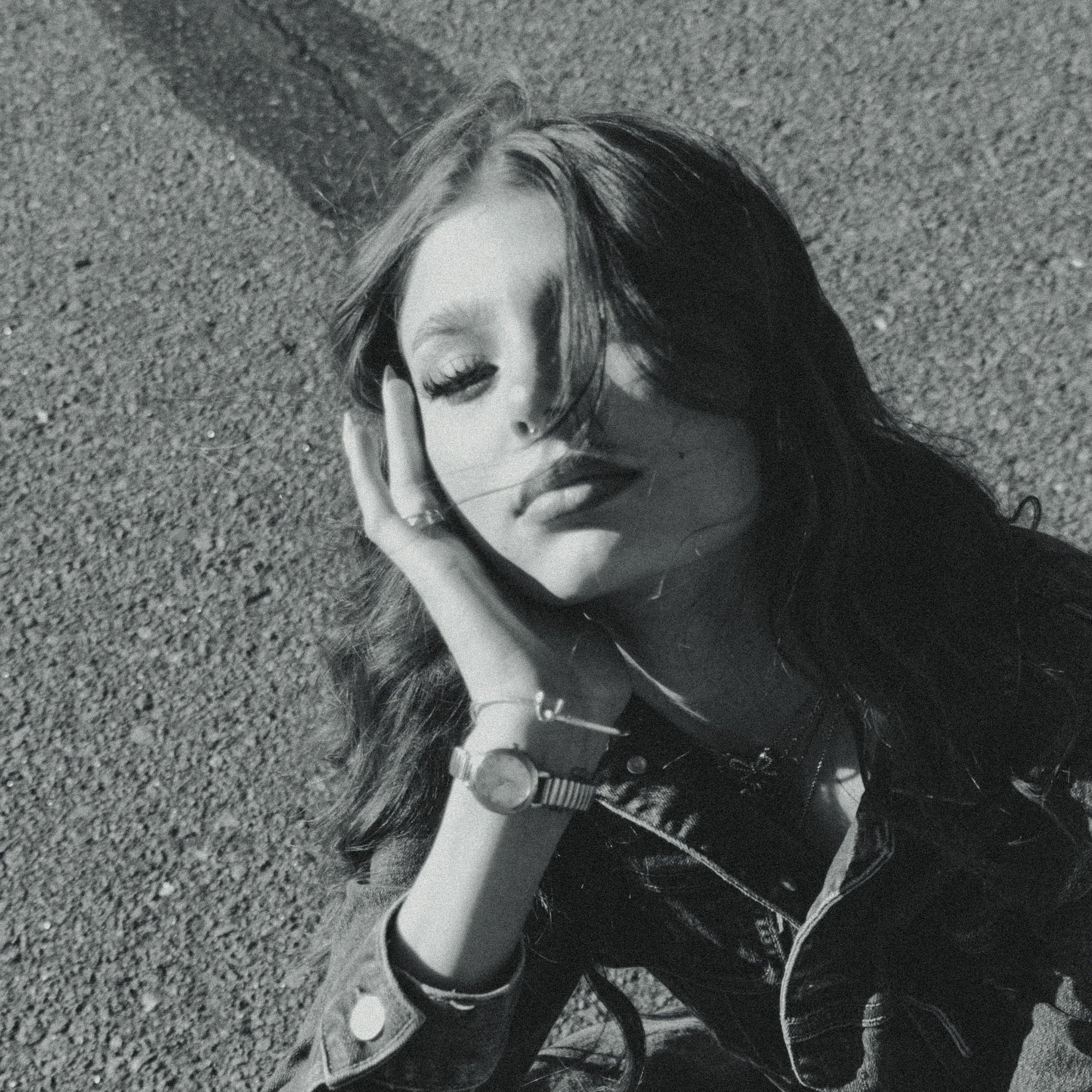 A black and white photo of a young woman with long hair, her eyes closed, resting her head on her hand, outdoors on a gravel surface, wearing a wristwatch and a jacket.