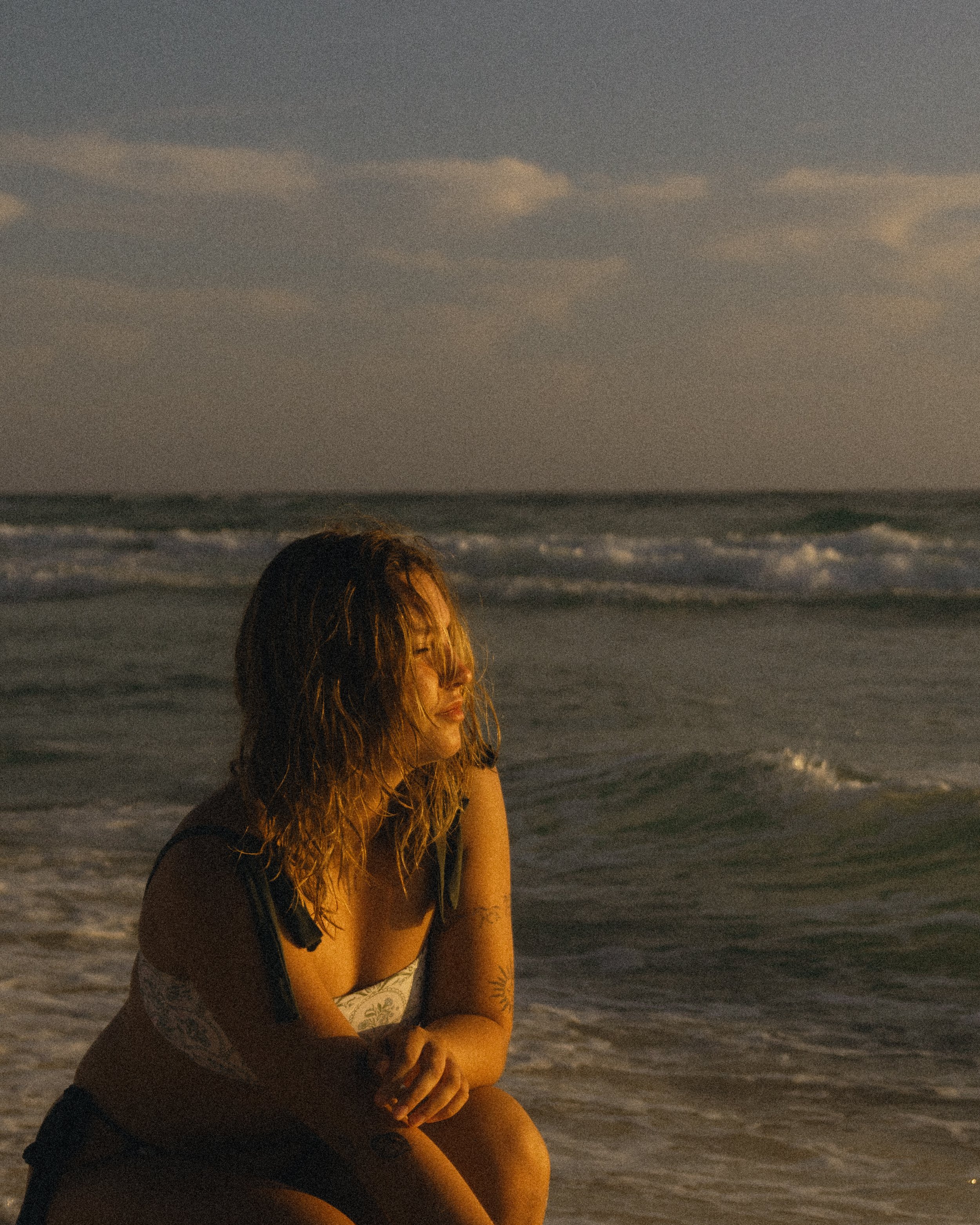 A woman with wet hair sitting on the beach at sunset, looking contemplative.