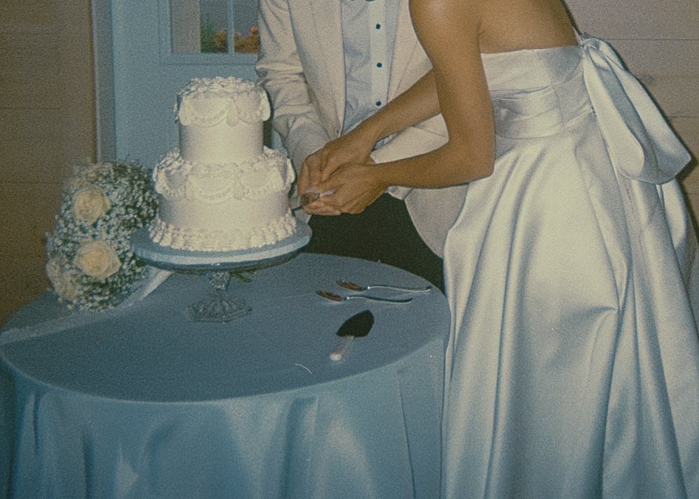 Bride and groom cutting their wedding cake, with bridal gown and flowers on the table.