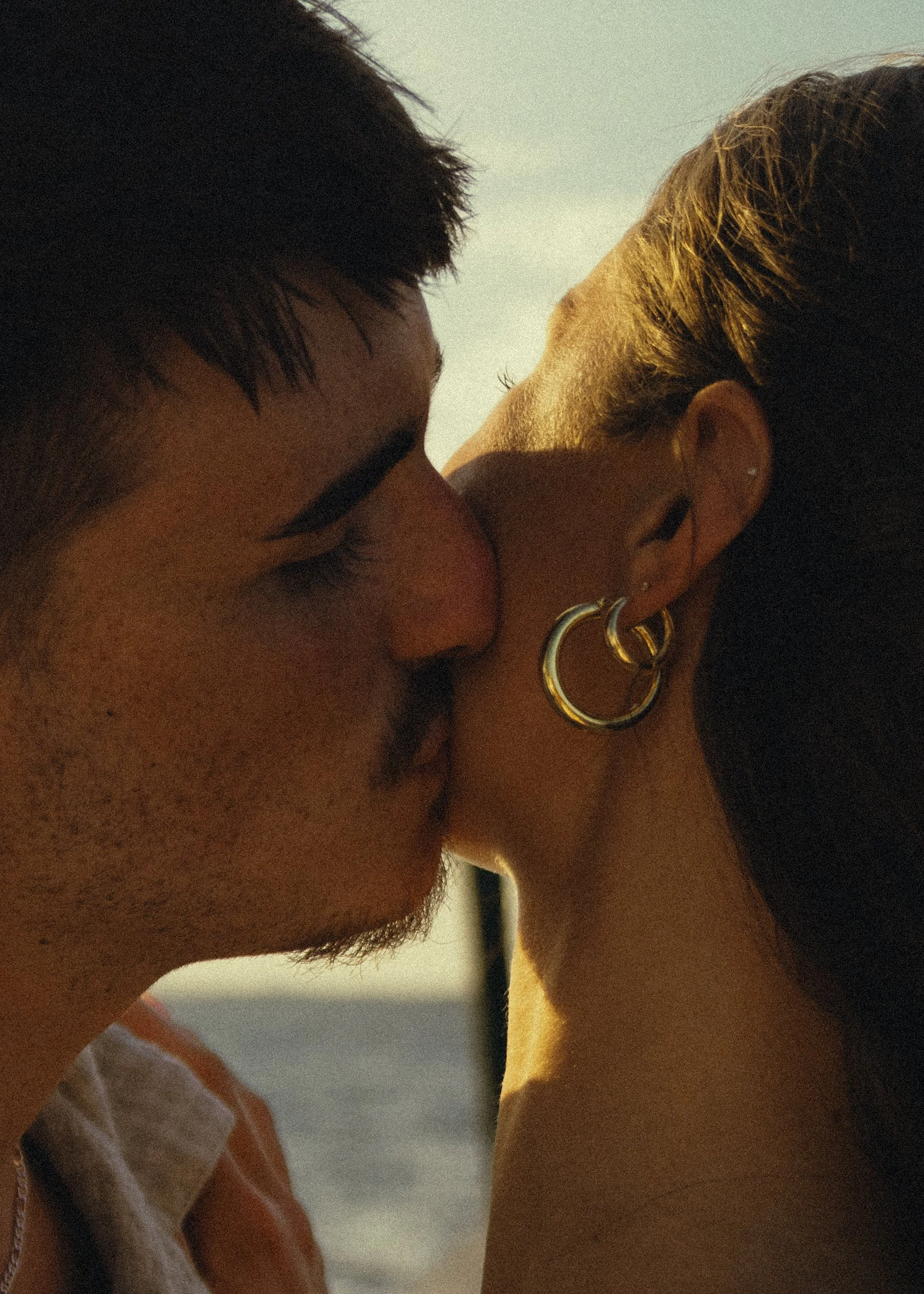 A close-up of a couple sharing a kiss outdoors, with the man's face on the left and the woman's ear and earrings visible on the right.