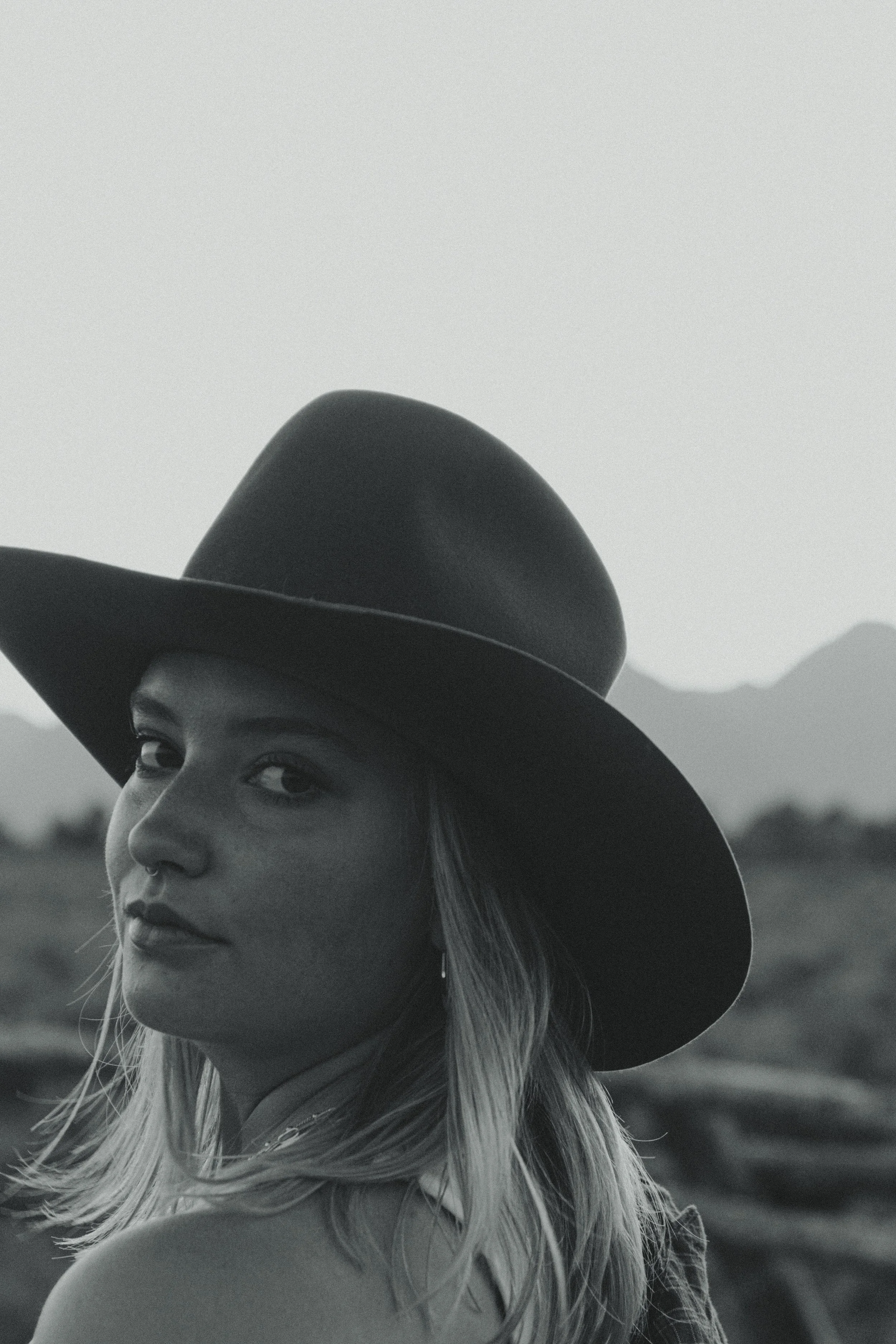 A black and white photograph of a woman wearing a wide-brimmed hat, looking over her shoulder with mountains in the background.