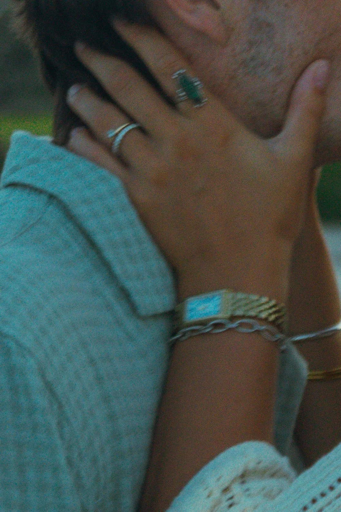 A person touching their face with their hand, wearing rings, a watch, and bracelets, and a gray hoodie.