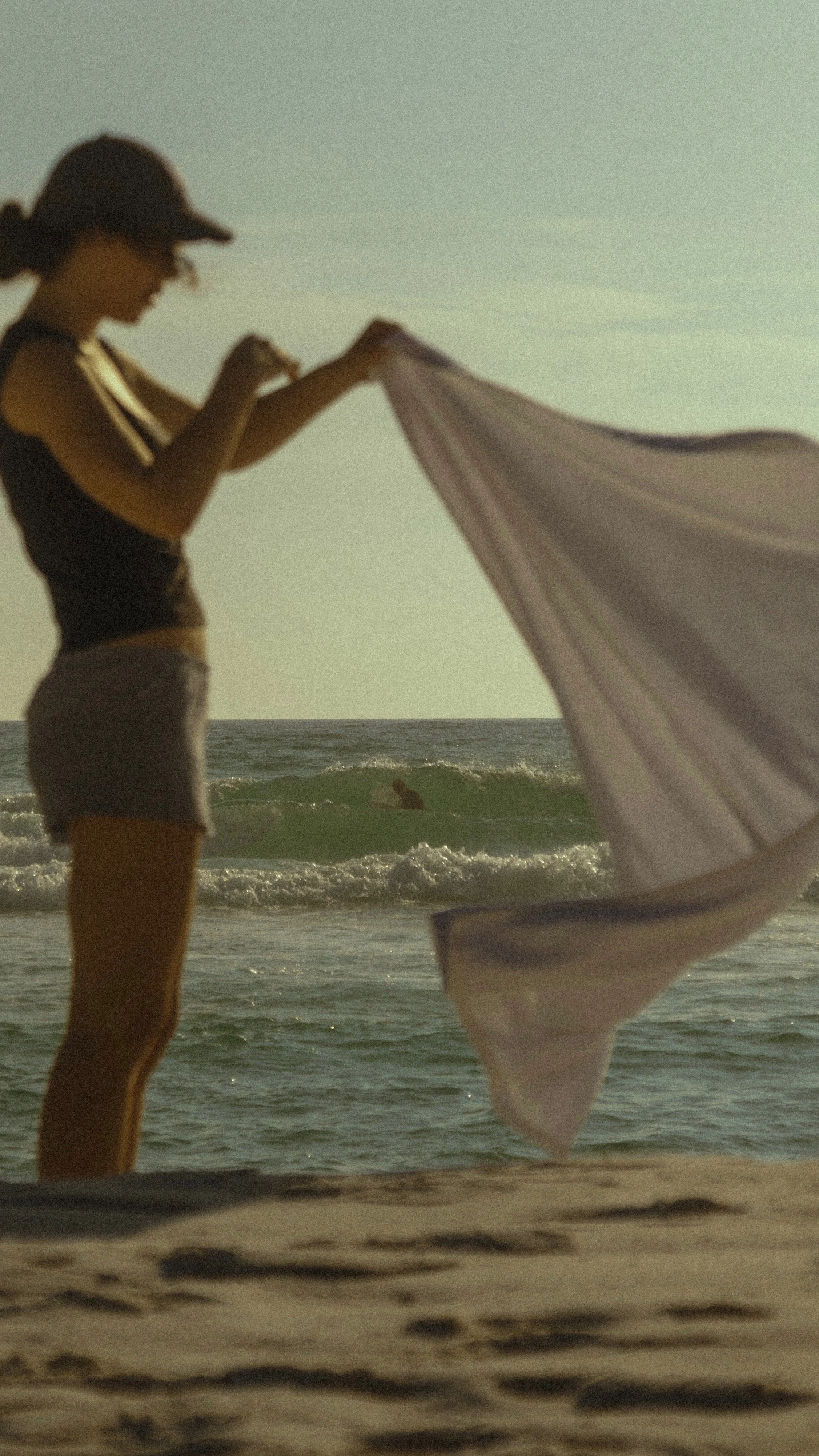A woman on the beach holding a beach towel or blanket, with ocean waves and a person surfing in the background