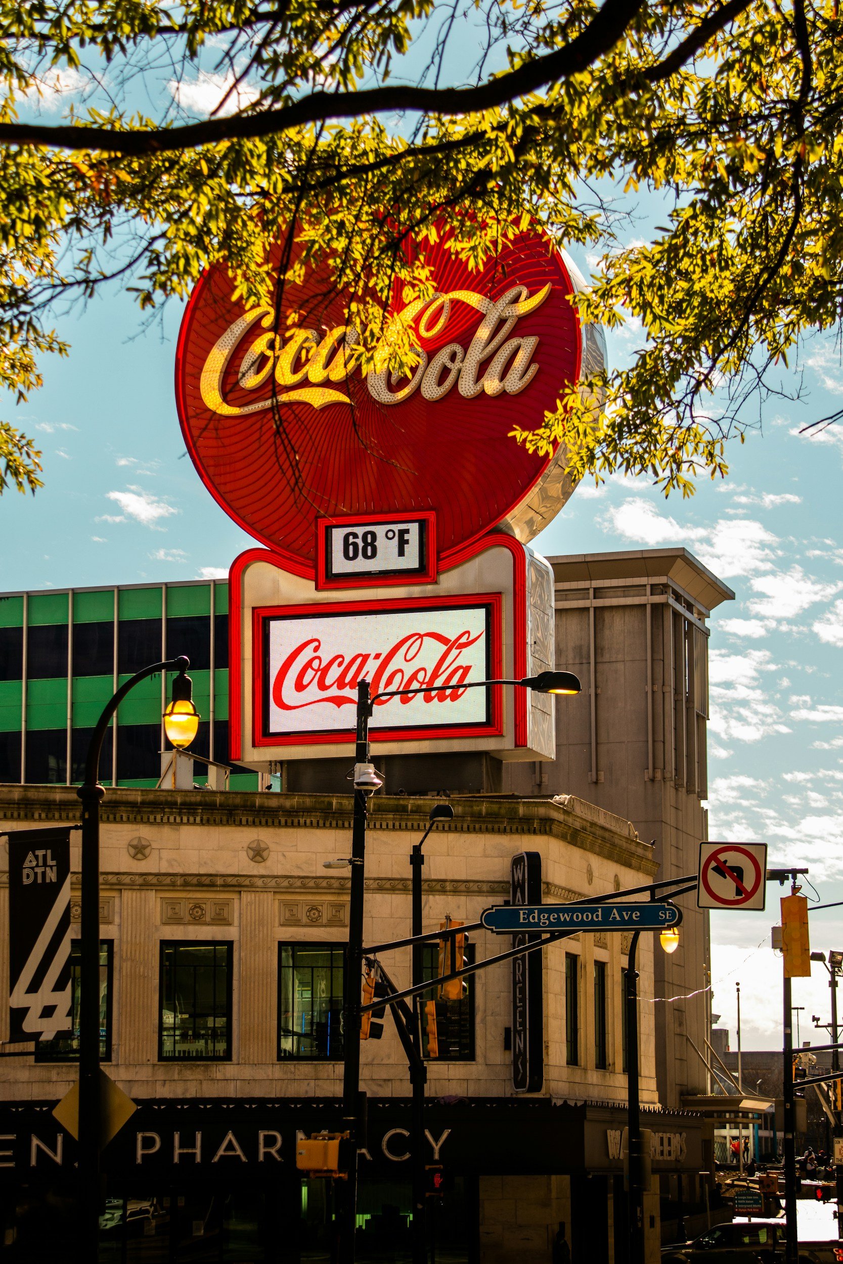 Large illuminated Coca-Cola sign above a city street, with a temperature display of 68°F, street signs for Edgewood Ave SE, and various streetlights and storefronts visible.