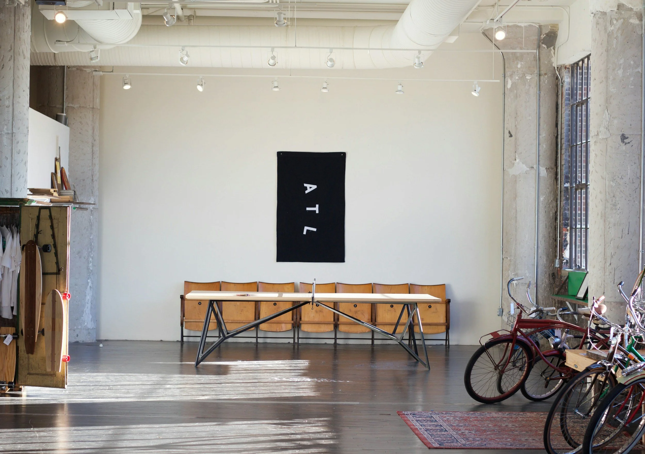 Interior of a modern, industrial-style room with a ping pong table, wooden chairs, bicycles, a rug, and a black wall hanging with white letters spelling "ATL."