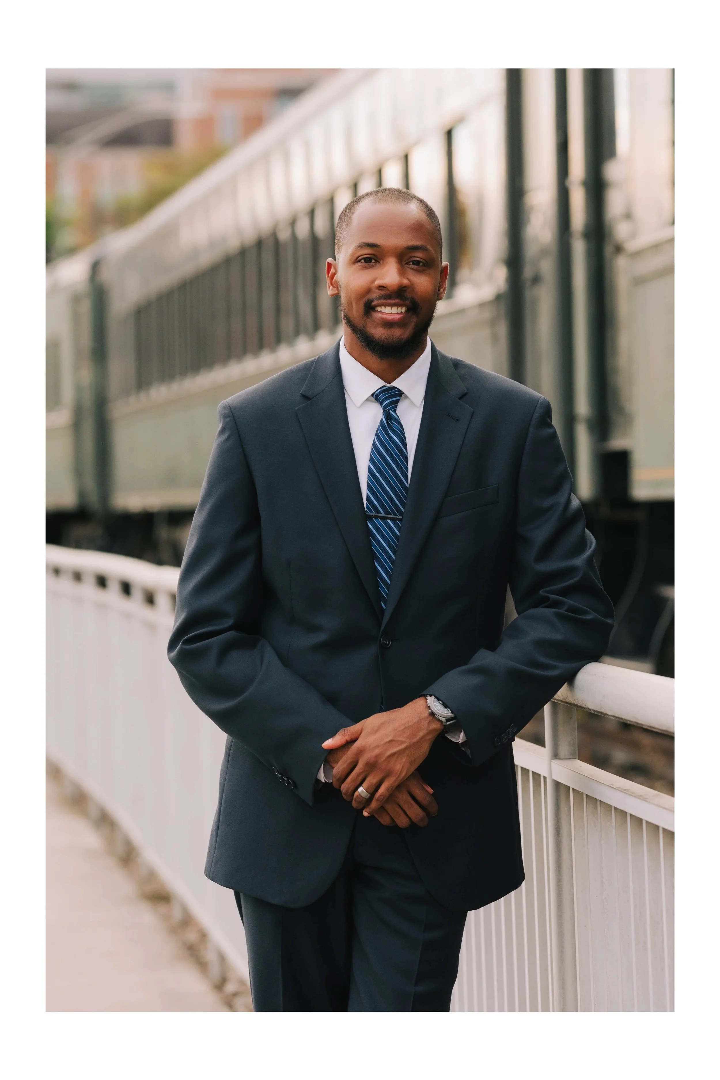 A man in a dark suit and striped tie standing outdoors on a bridge near a train, smiling at the camera.