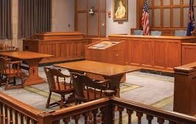 Empty courtroom with wooden benches, judge's bench, American flags, and an American flag in the background.