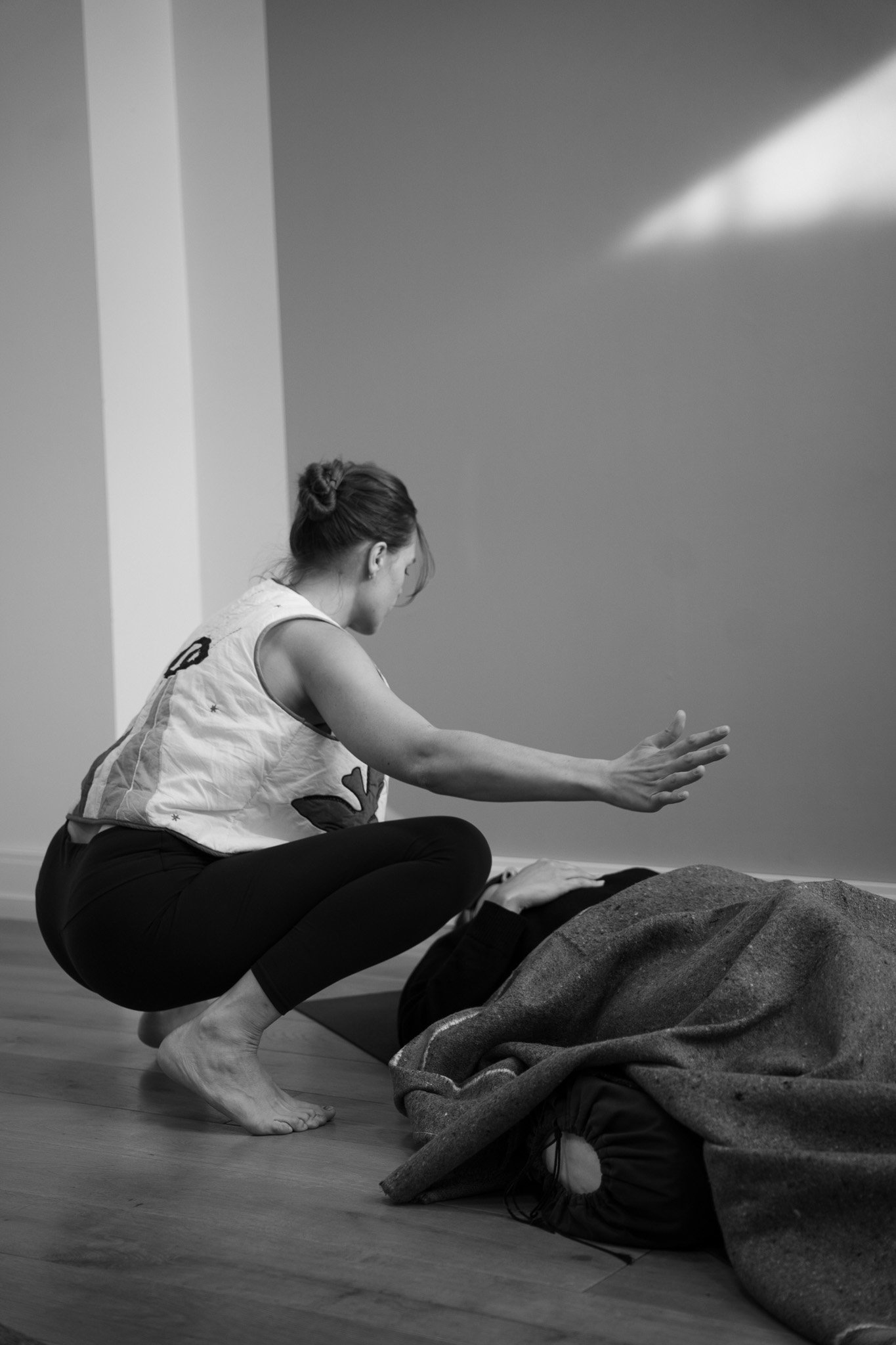 Une femme guide une séance de breathwork sur une personne allongée couverte d'une couverture dans une pièce intérieure. La femme est accroupie, les bras étendus en avant, dans une posture attentive.