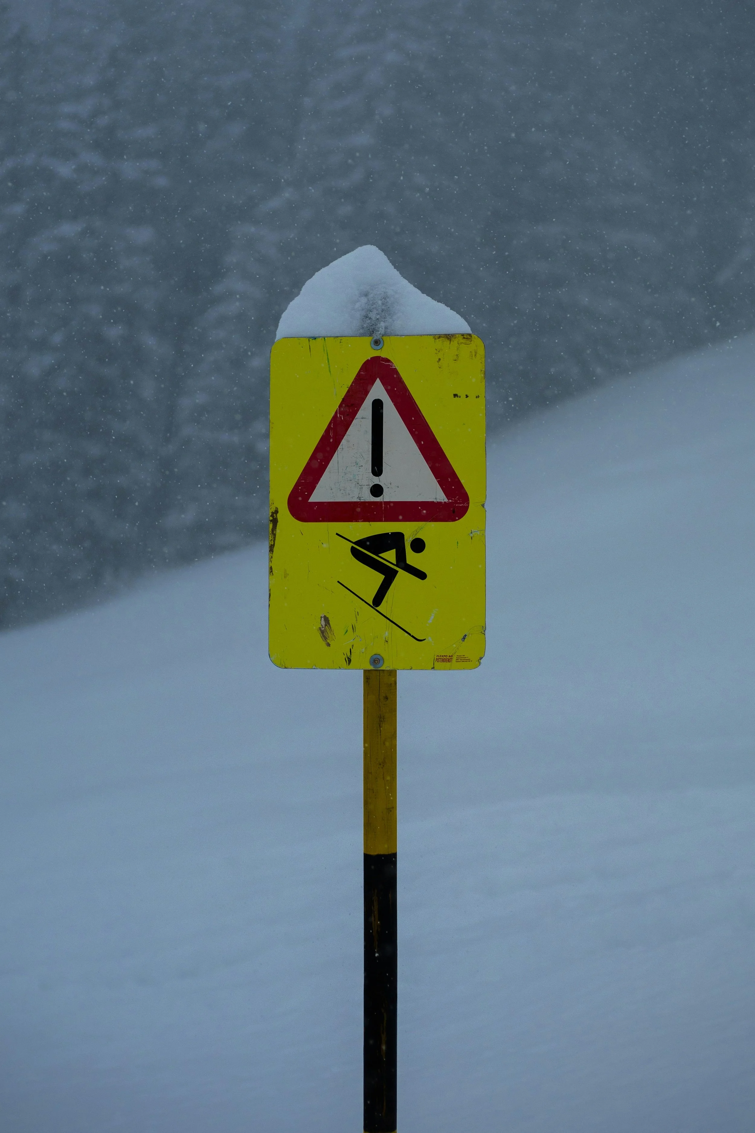 A warning sign covered with snow in a snowy landscape with trees in the background.