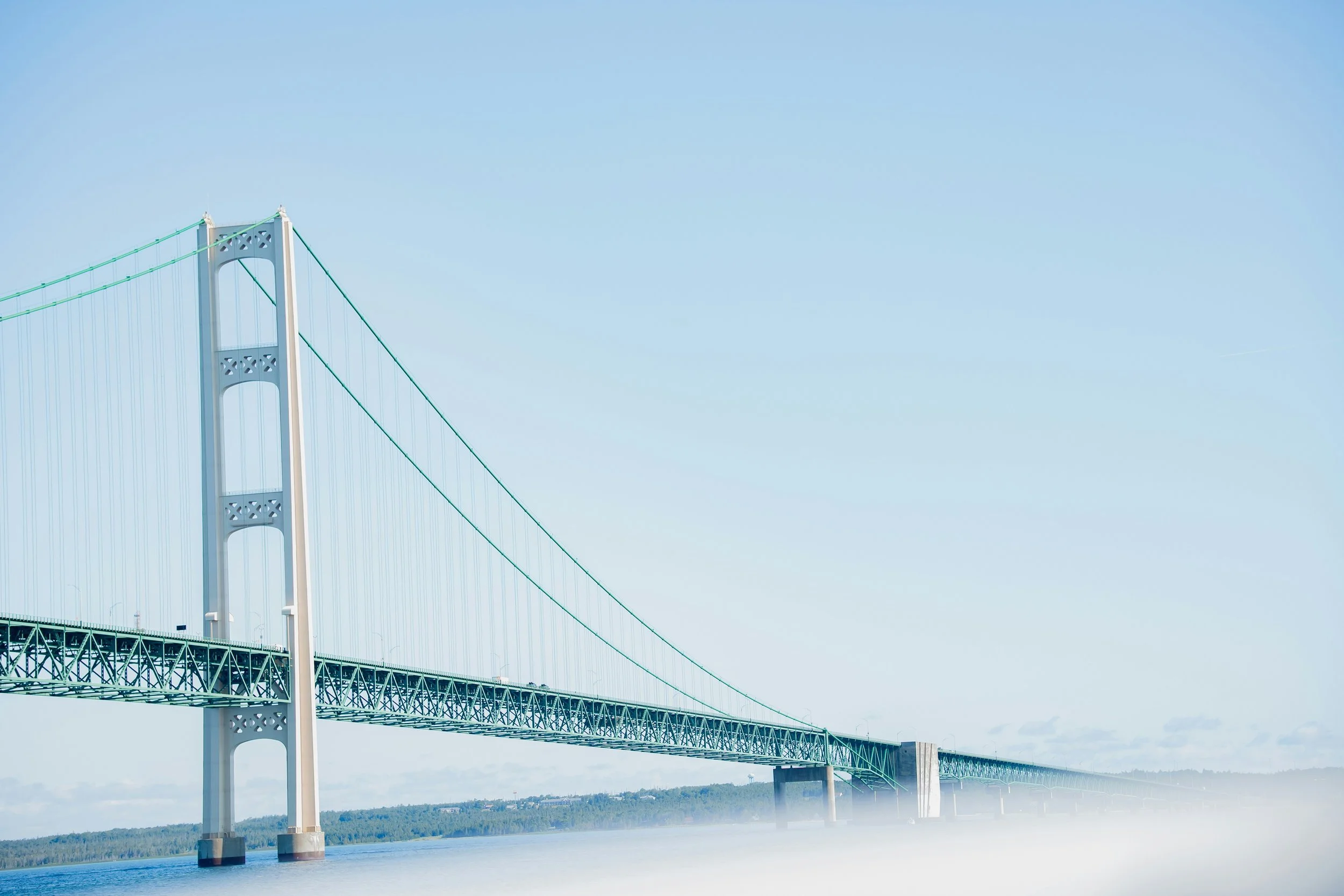 A large suspension bridge extending over a body of water with hills in the background, under a clear blue sky. Mackinac Bridge