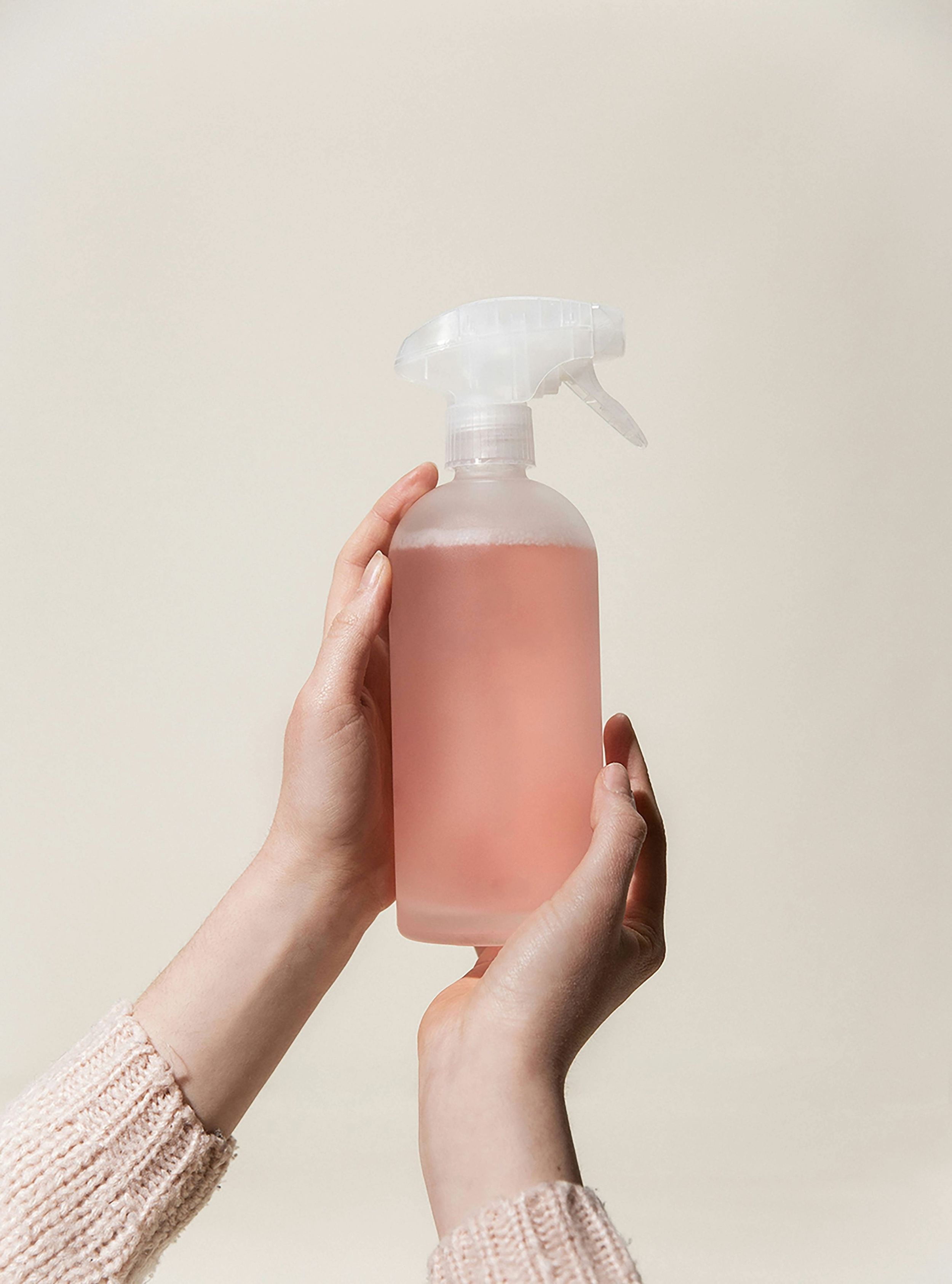 A person holding a spray bottle filled with pink liquid soap or cleaning solution against a plain background.